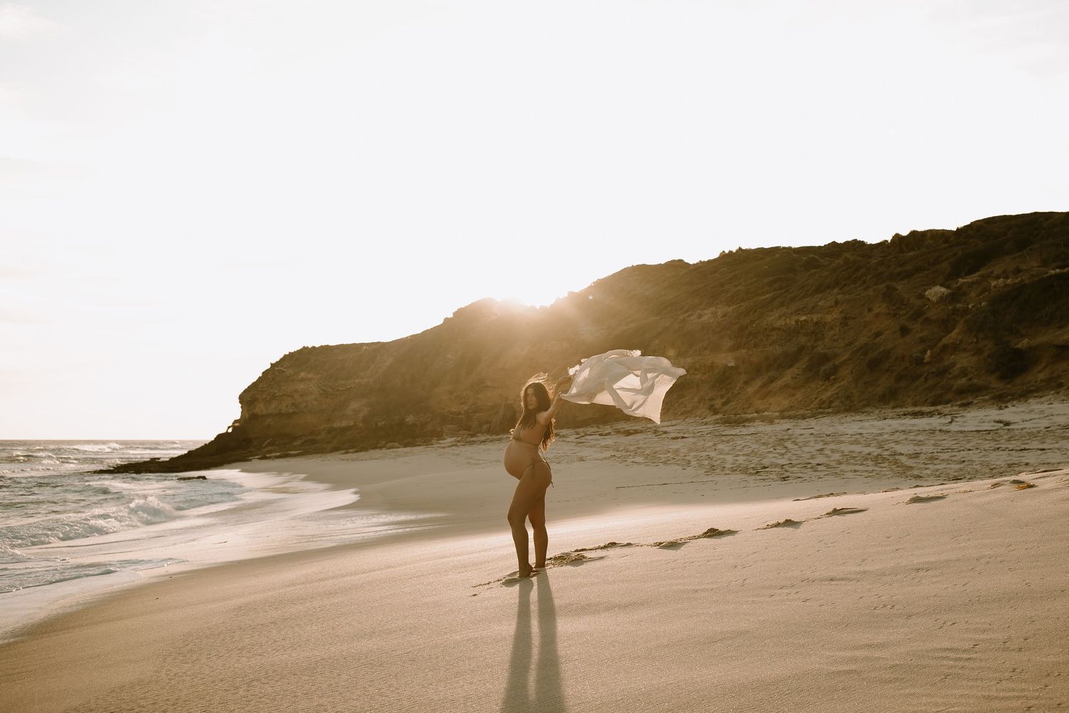 Silhouette of a person holding flowing fabric on a beach at sunset with cliffs in the background.