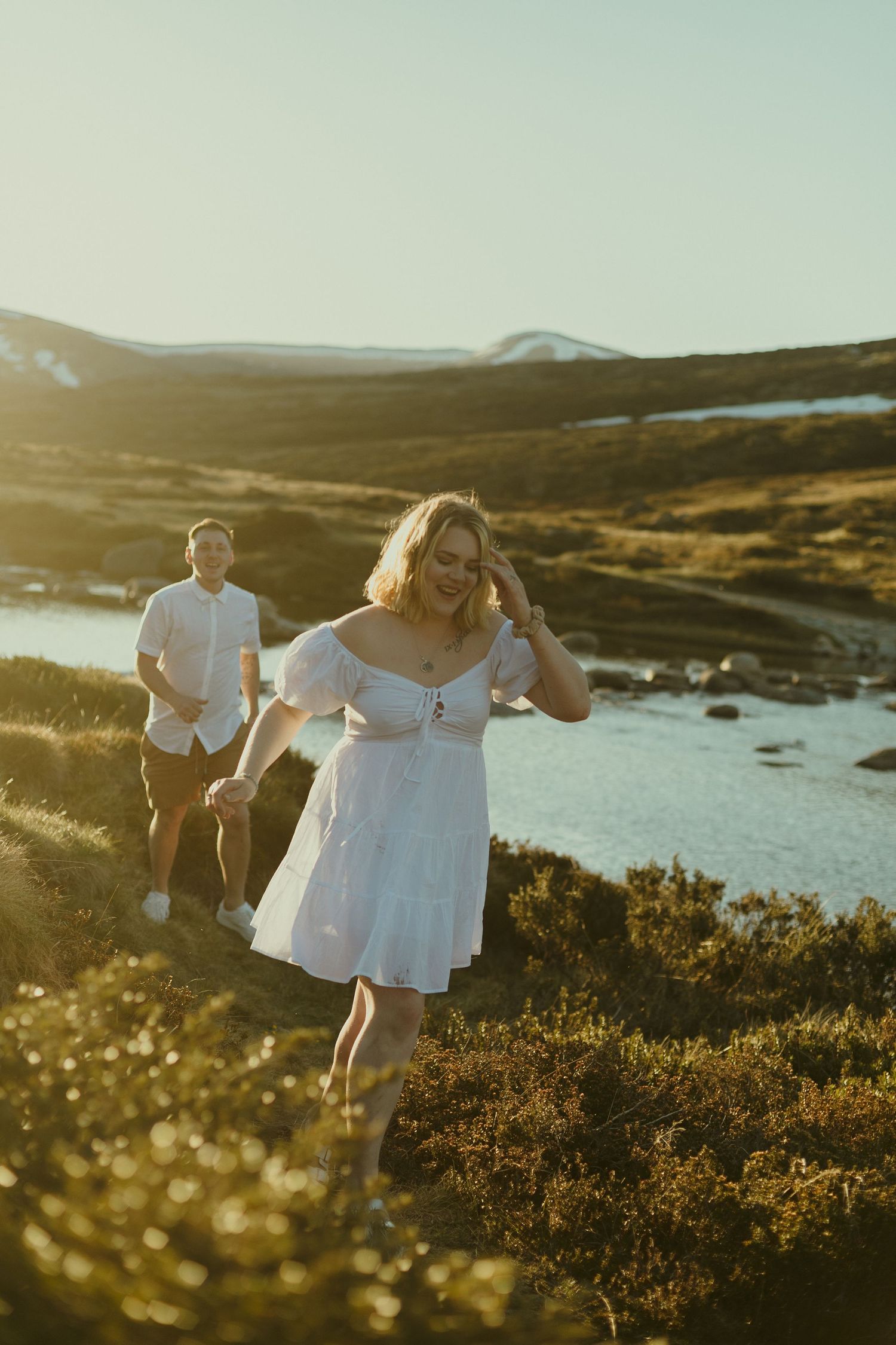 A couple walks along a scenic coastal hillside path at golden hour with mountains and water in the background.