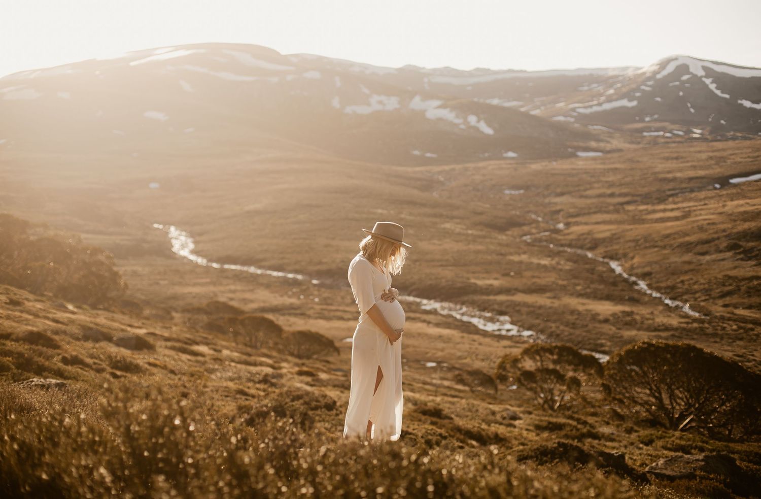A lone figure stands on a hilltop at sunset, overlooking a winding river through golden mountain valleys.