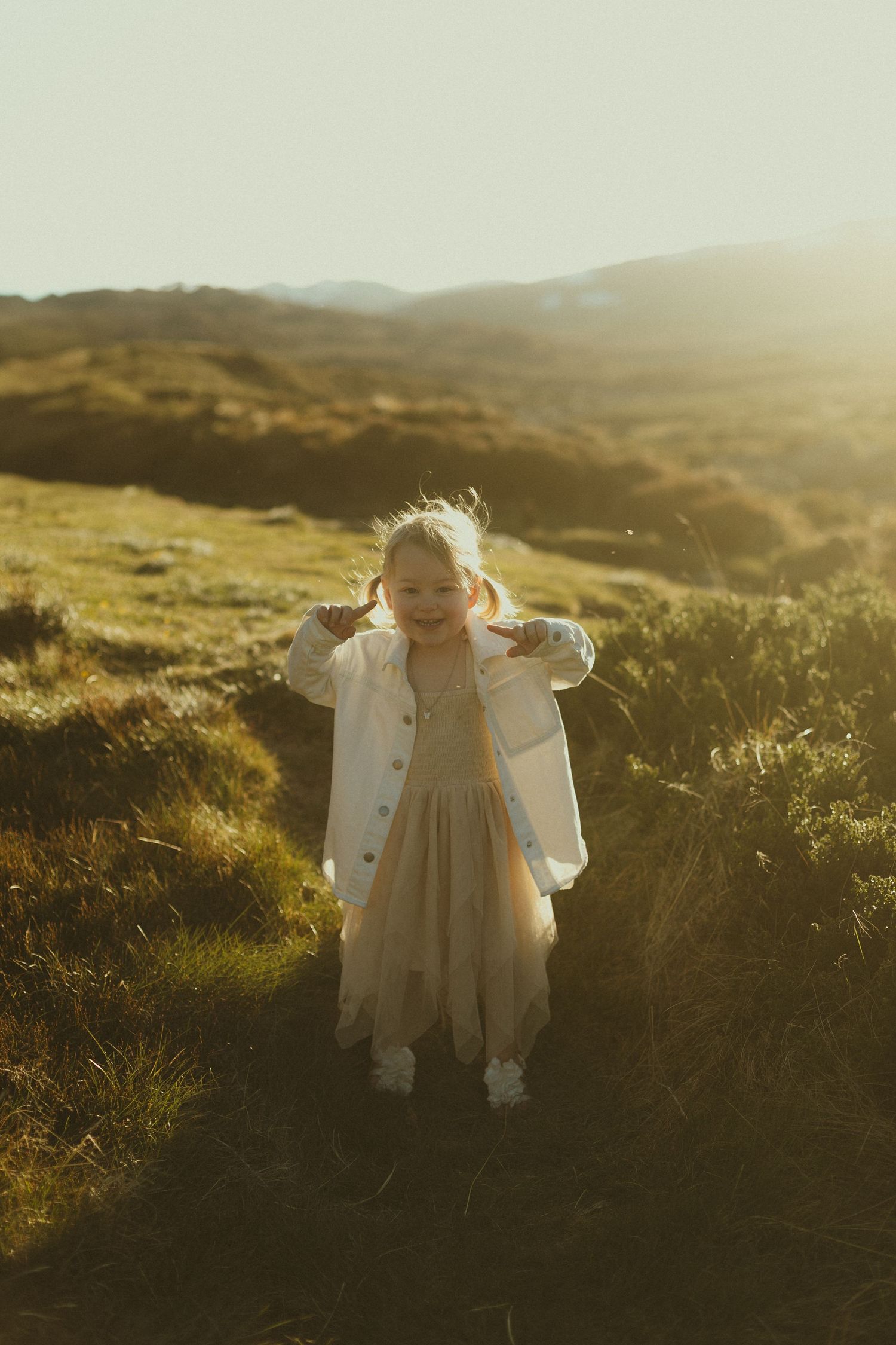 A dreamy golden hour photo on a grassy hillside features a white dress and jacket ensemble against a mountainous backdrop.