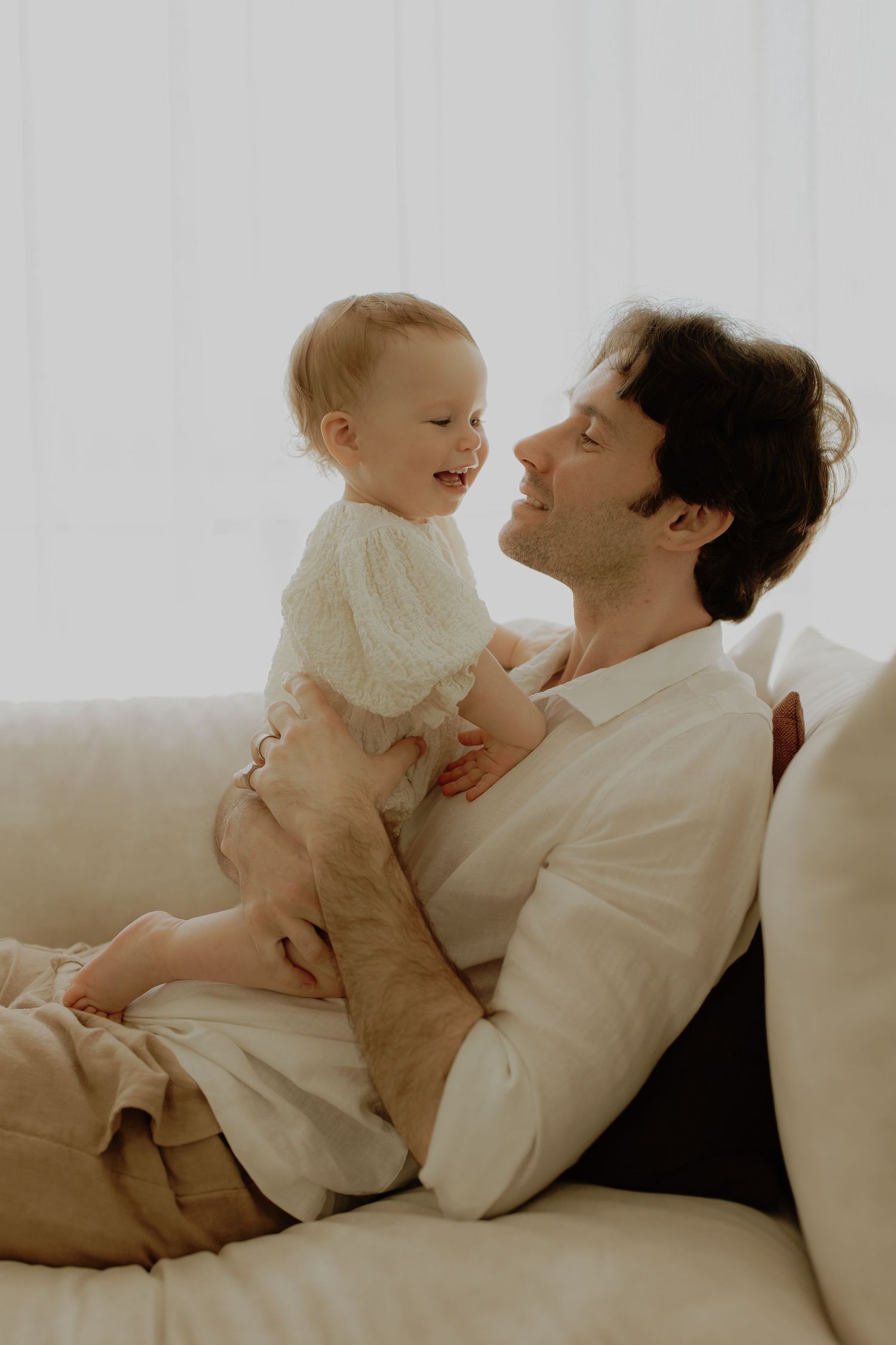 Father relaxing on a white sofa, lovingly holding his smiling baby dressed in a white outfit.