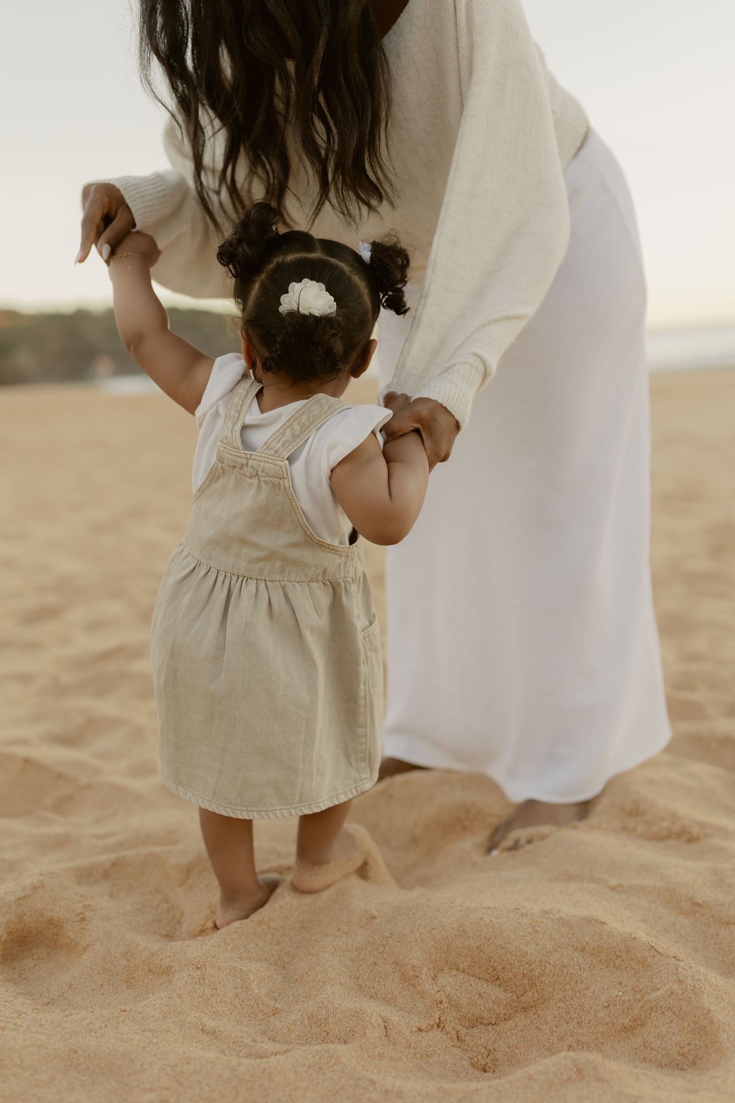 Mother in white dress holding toddler daughters hands while walking barefoot on sandy beach.
