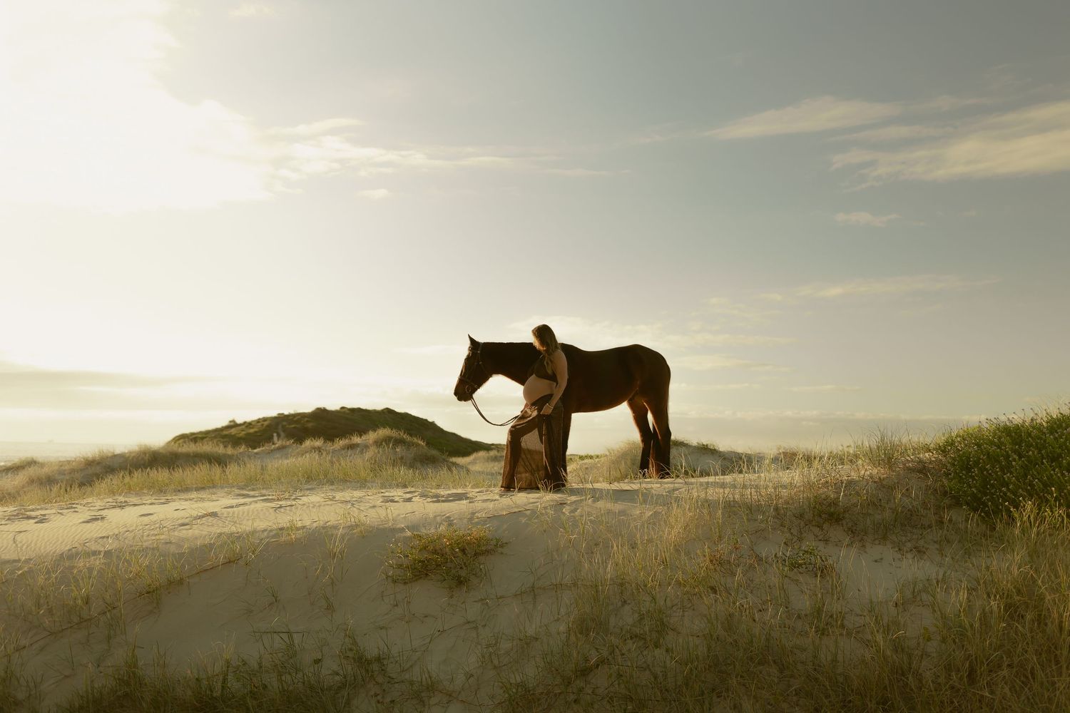 A person stands with a brown horse on sandy dunes with grass under a bright sky.