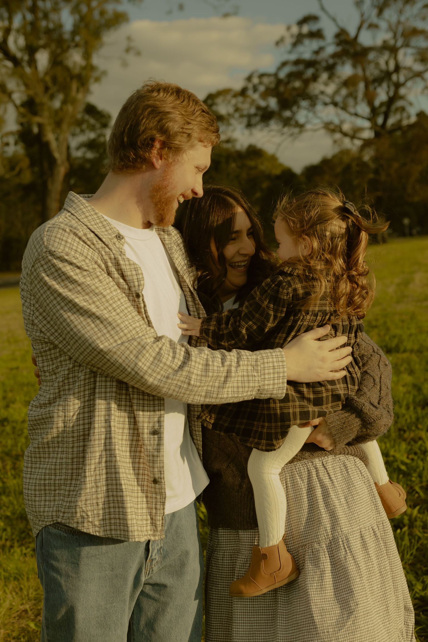 A young family with a toddler girl shares a warm embrace in a sunlit green field during golden hour.