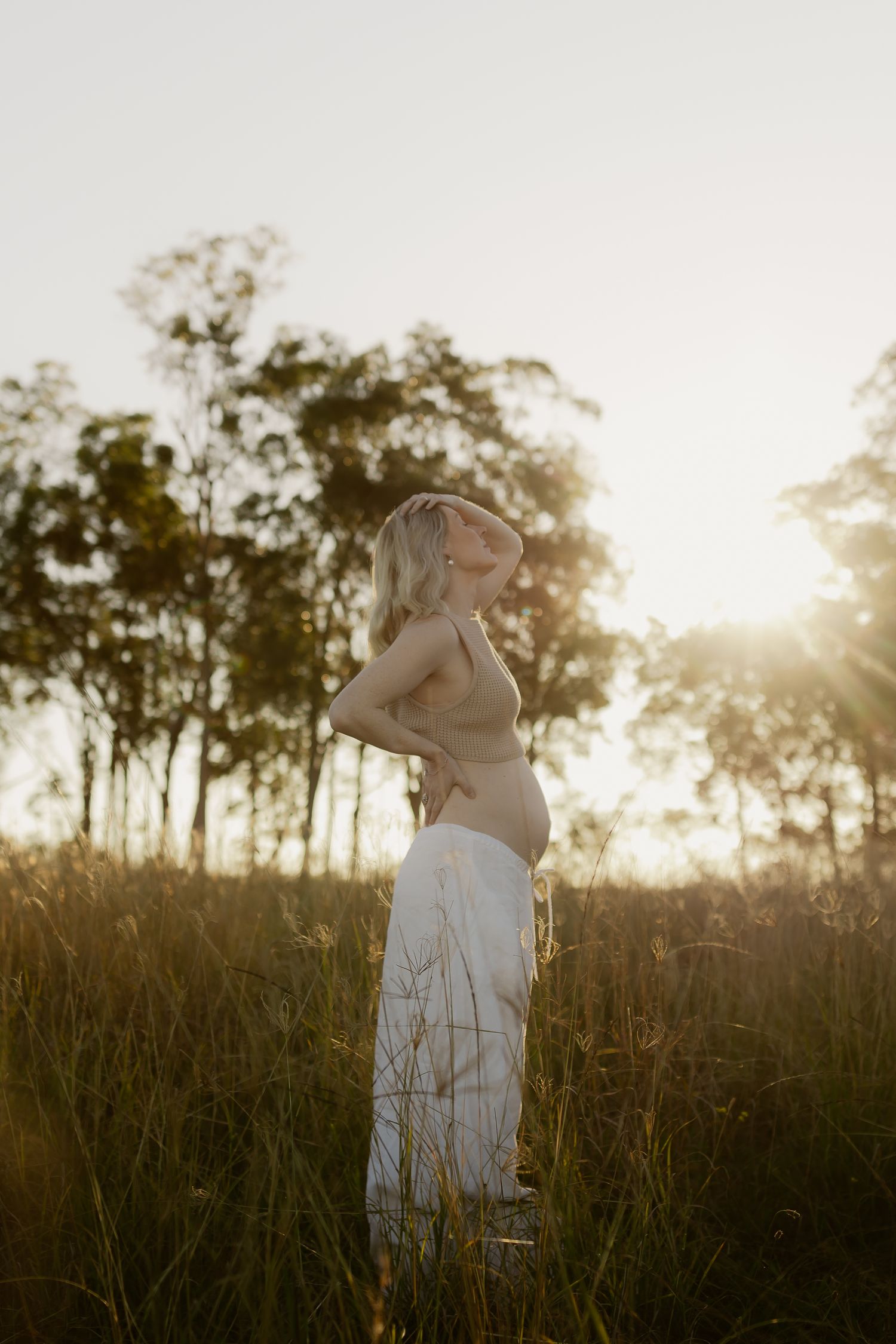 Pregnant woman in white skirt standing in golden field at sunset with trees in background.
