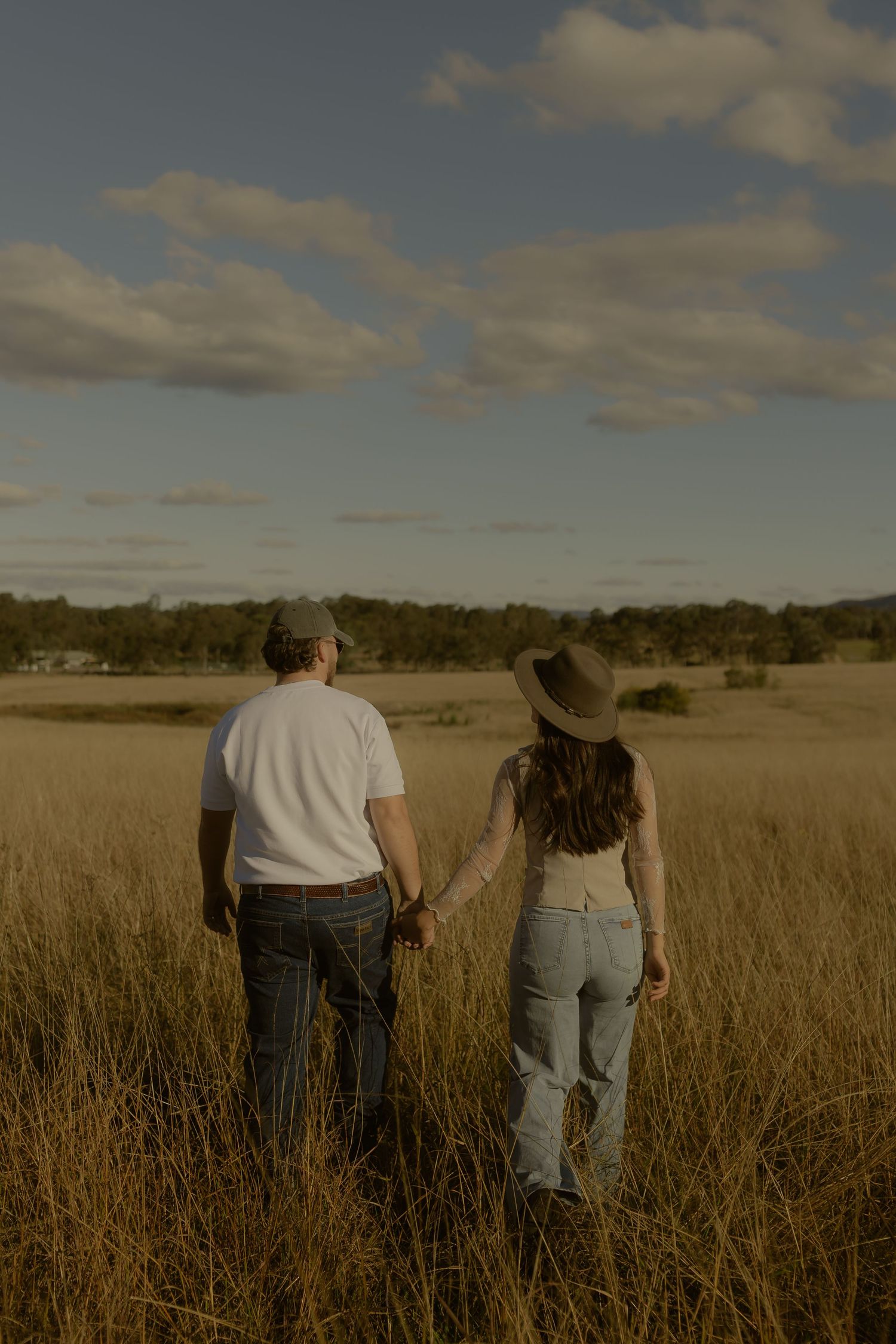 Couple holding hands walking through golden grass field under cloudy sky, viewed from behind.
