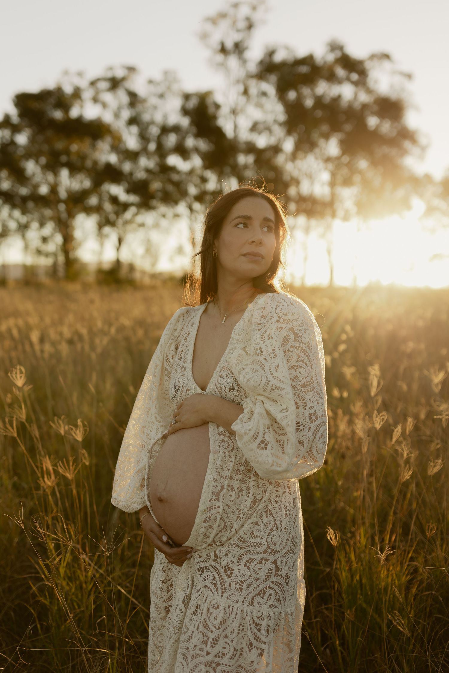 Pregnant woman in white lace dress cradling baby bump in sunlit field at golden hour.