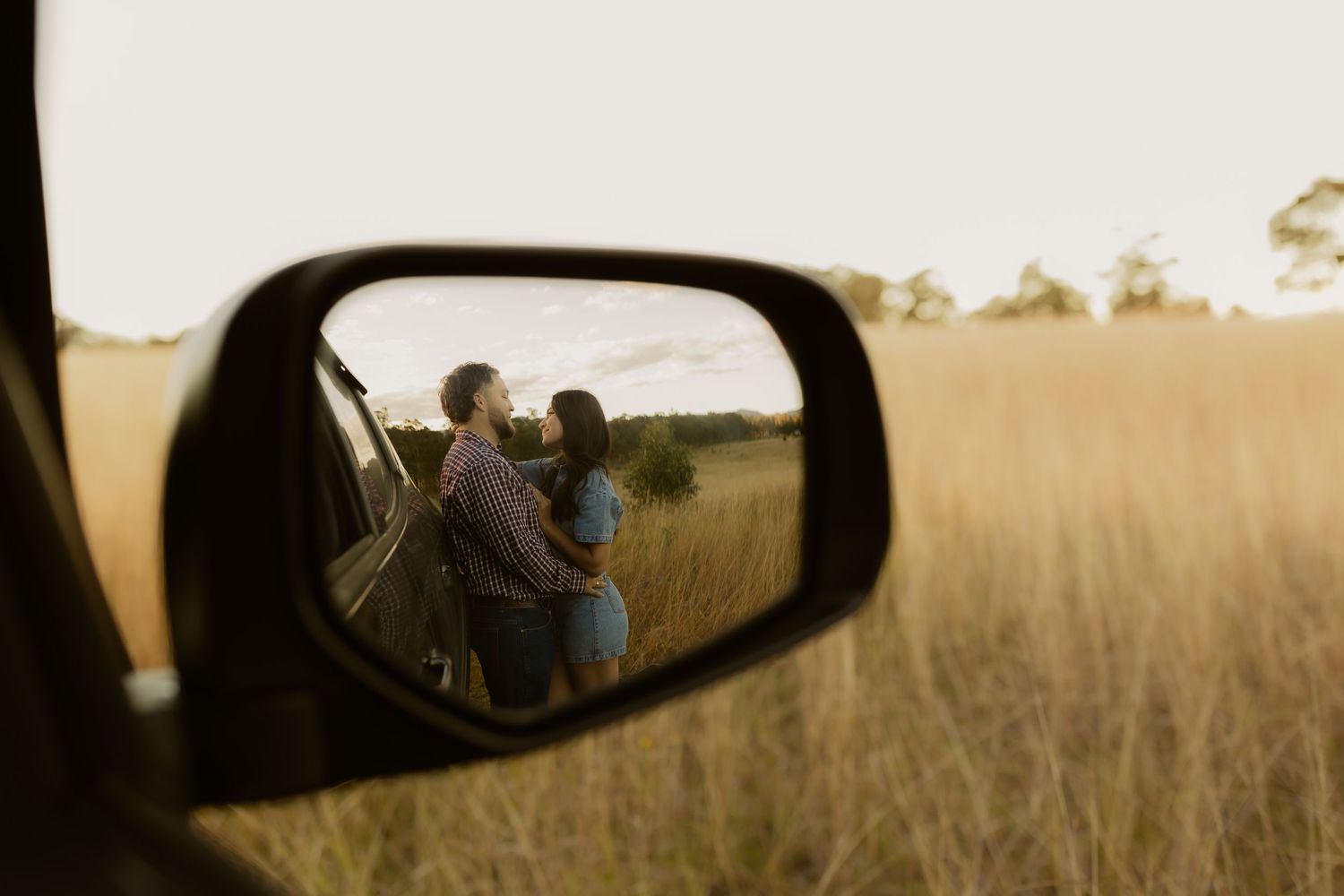 A couple embraces in a golden field, reflected in a car side mirror at sunset.
