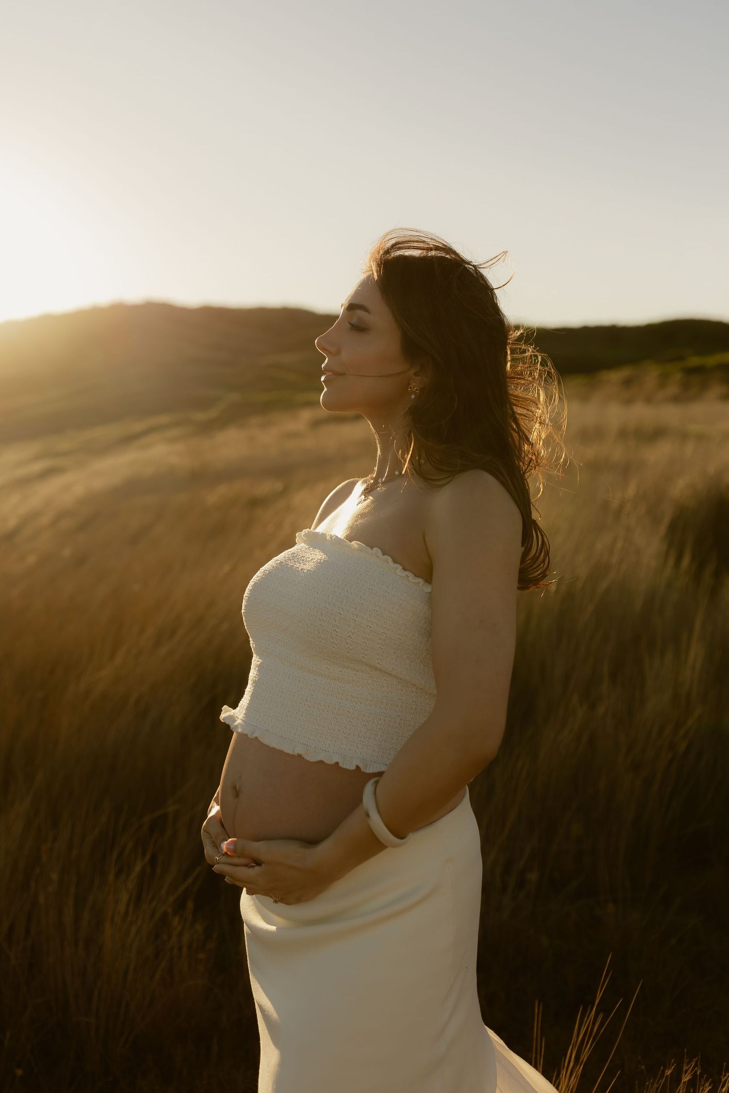 Pregnant woman in white strapless dress cradling her baby bump at golden hour in a field of tall grass.