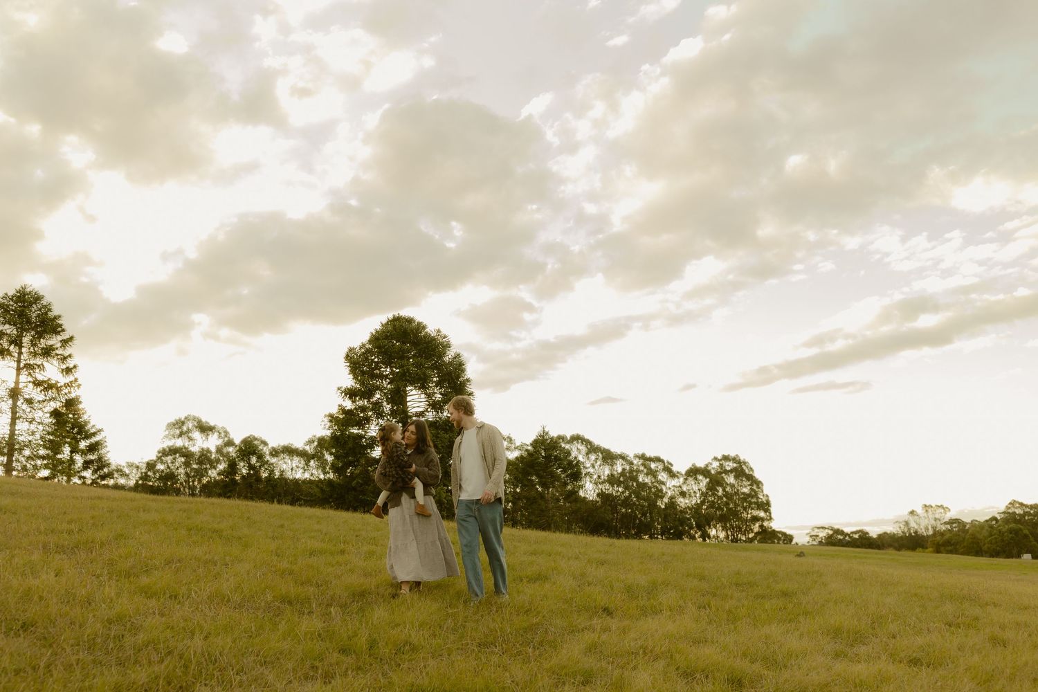 Couple walking hand-in-hand across a lush green field at golden hour with dramatic clouds overhead.