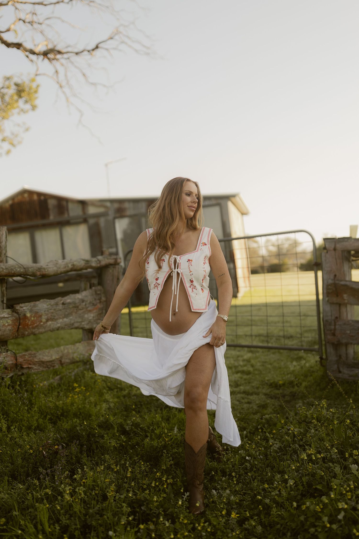 Woman in floral crop top and white skirt with cowboy boots poses near a farm gate at golden hour.