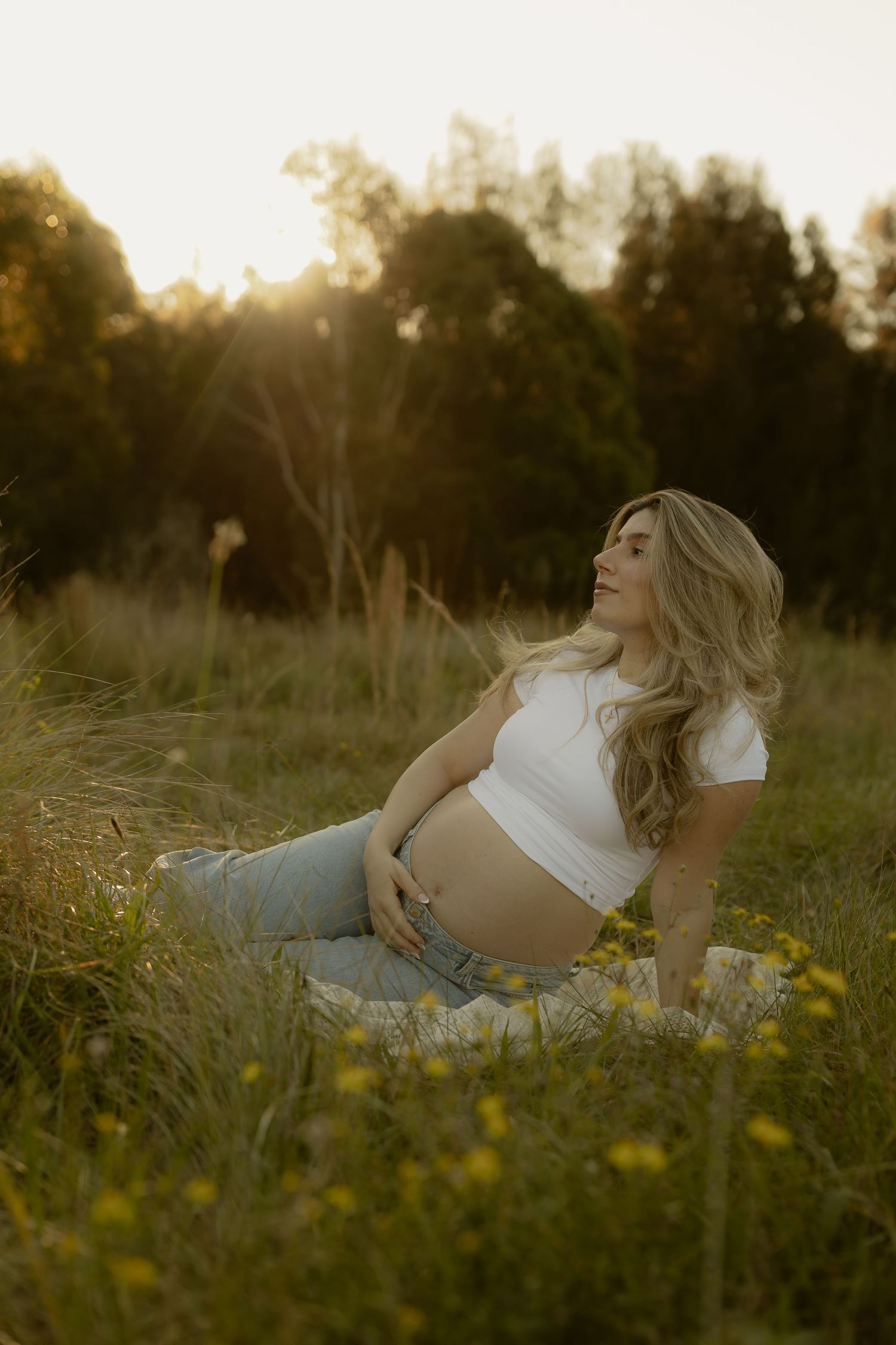 Pregnant woman in white crop top and jeans sitting in sunlit meadow with wildflowers, golden hour maternity photo.