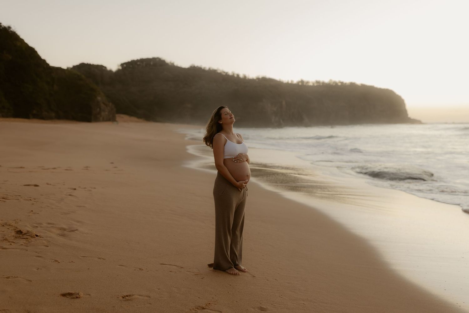 Pregnant woman in white top and olive pants standing on a sandy beach at golden hour sunset.