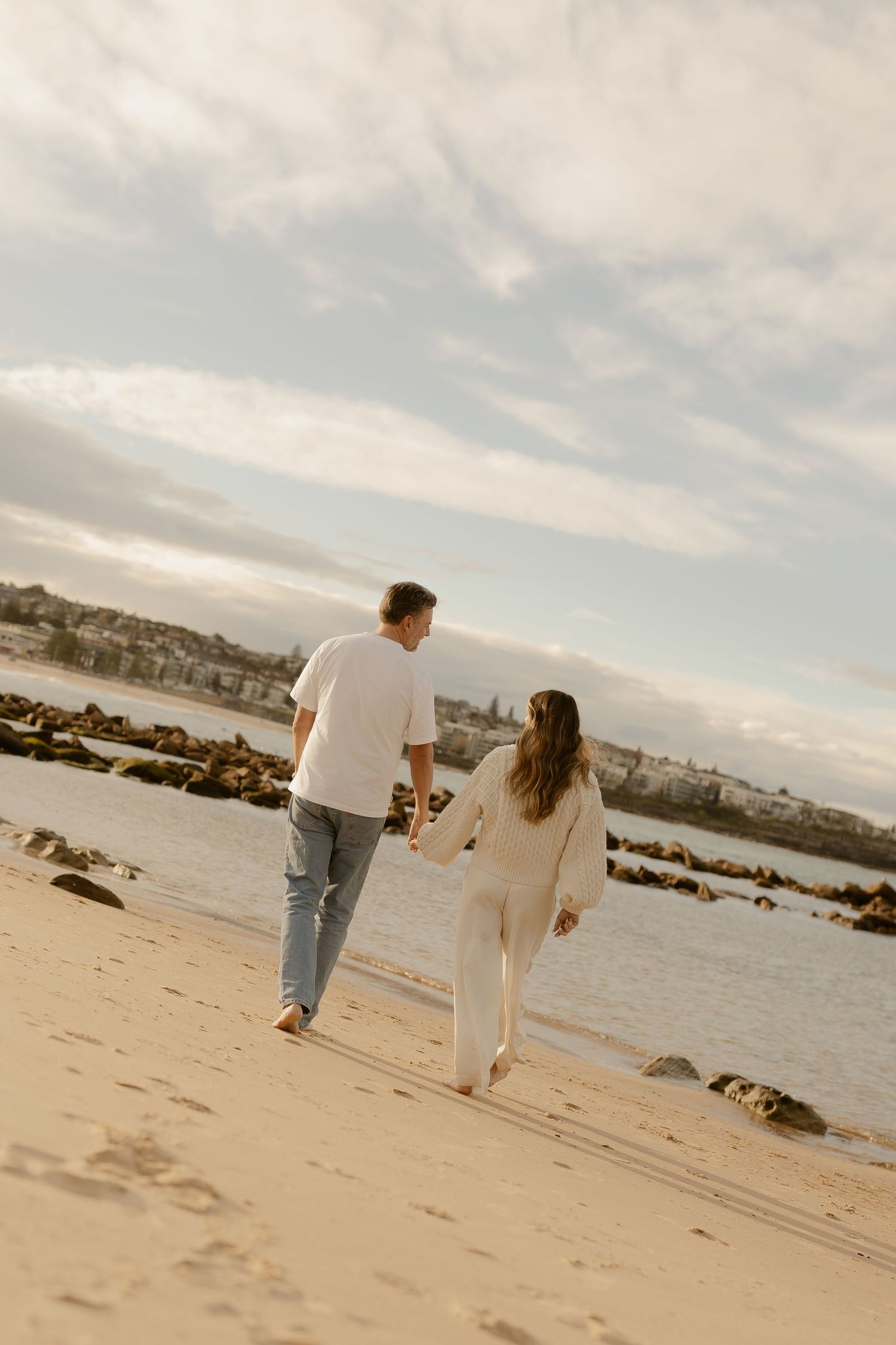 Couple holding hands walking on sandy beach at golden hour, wearing casual white outfits with ocean and rocky shore in background.