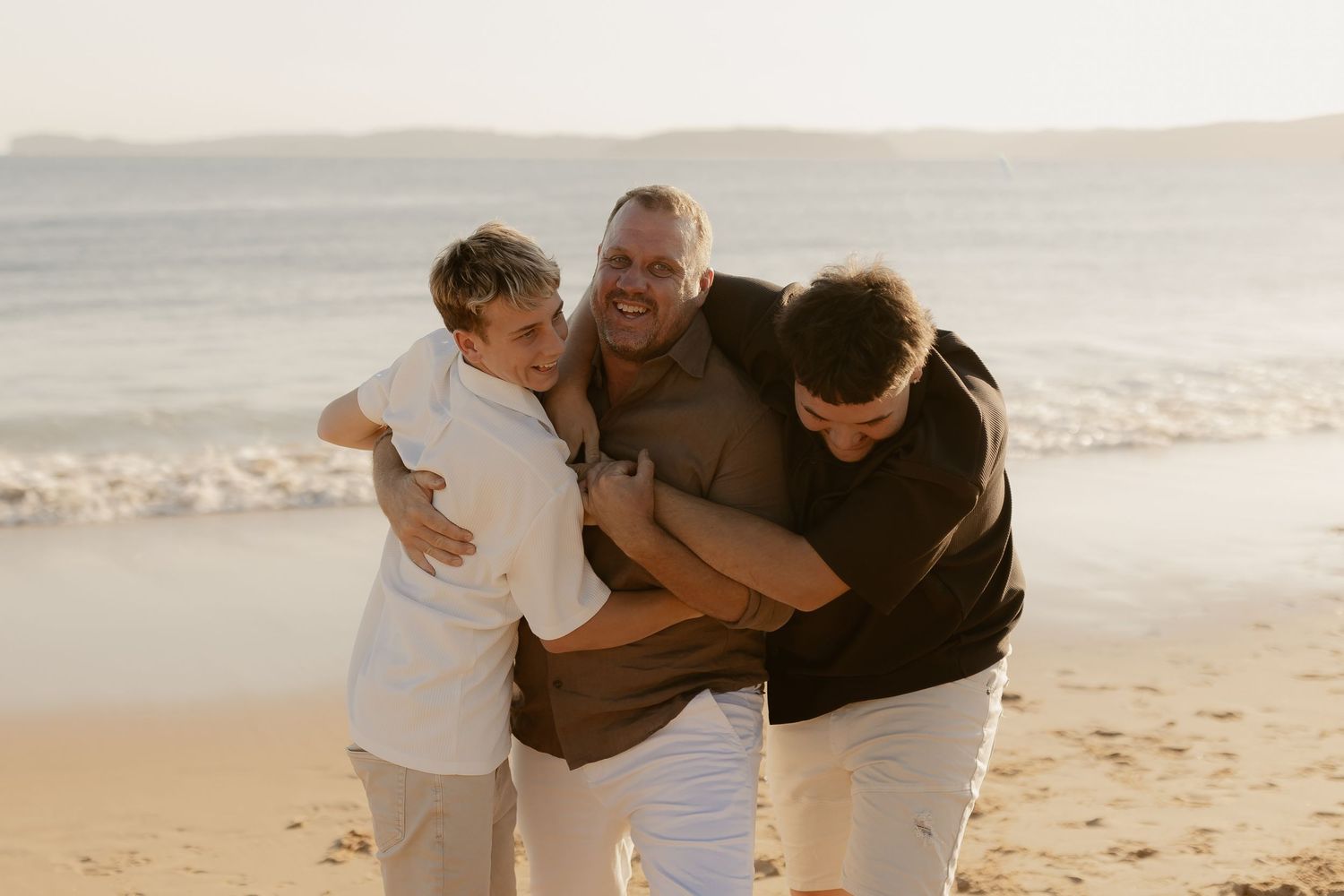 Three people embrace and laugh together on a sunny beach at golden hour.
