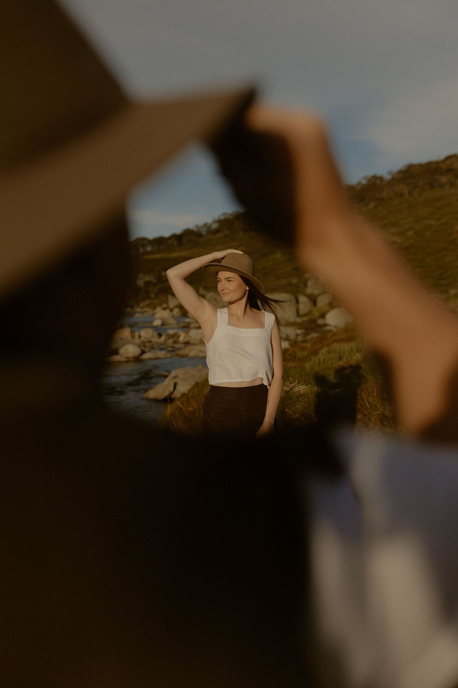 Person in a straw hat poses by a scenic lake surrounded by rocky terrain and greenery at sunset.