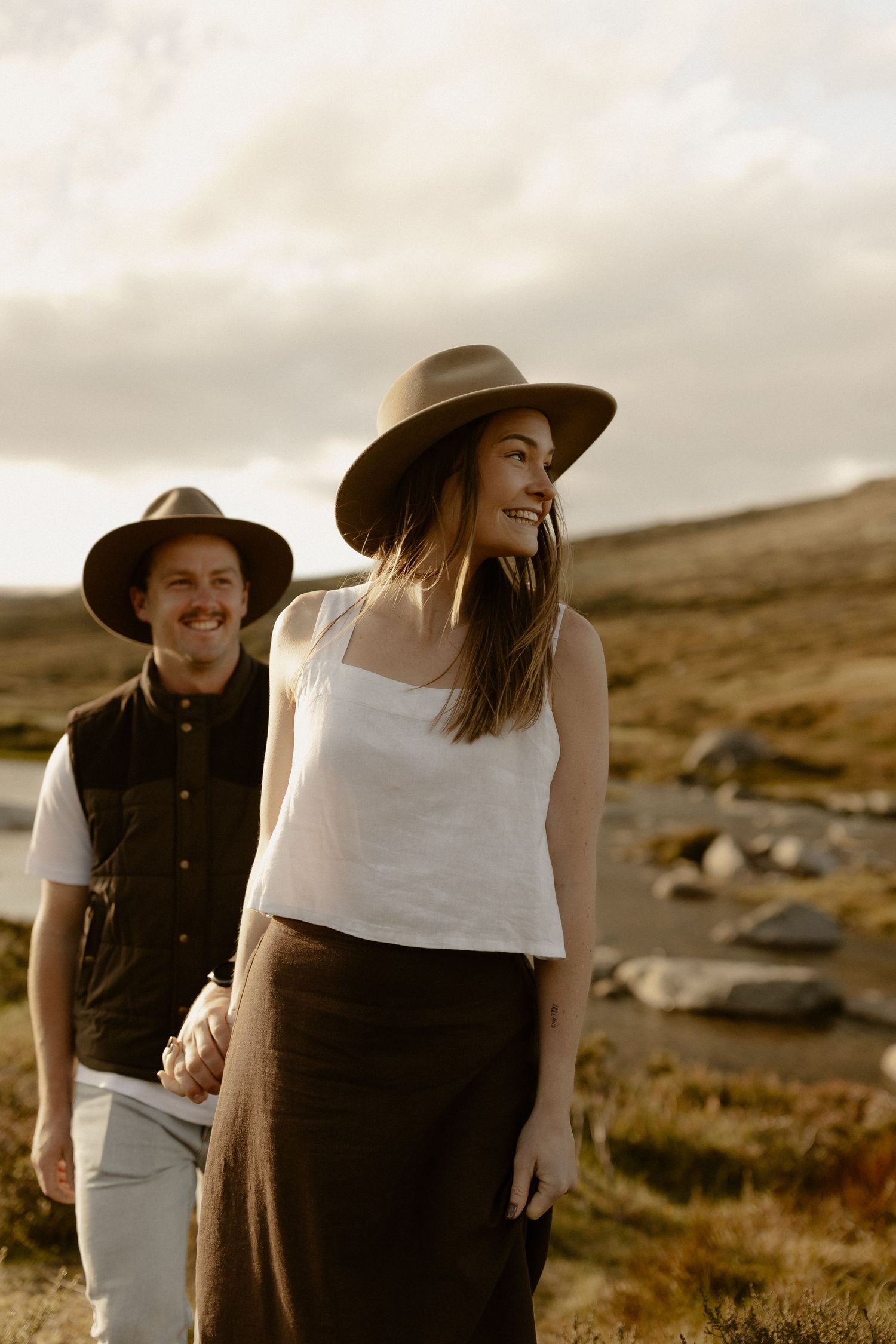 Couple in western attire poses together against a scenic mountainous landscape at sunset.