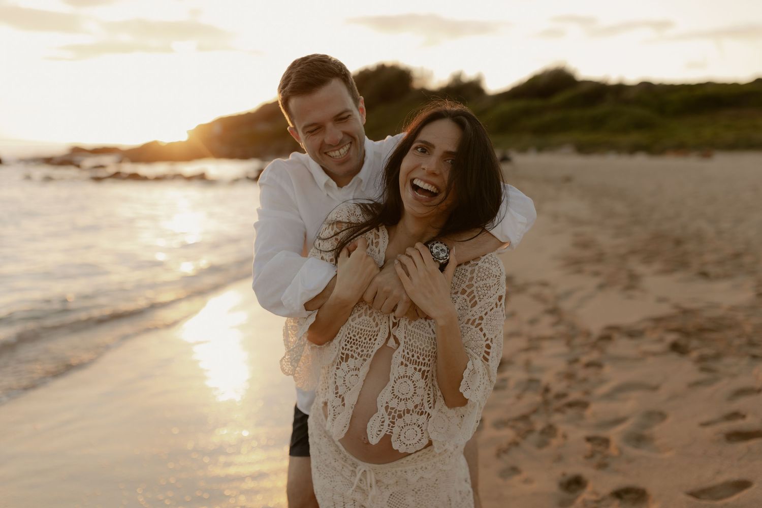 A joyful couple embraces on a sandy beach at sunset with ocean waves and cliffs in the background.