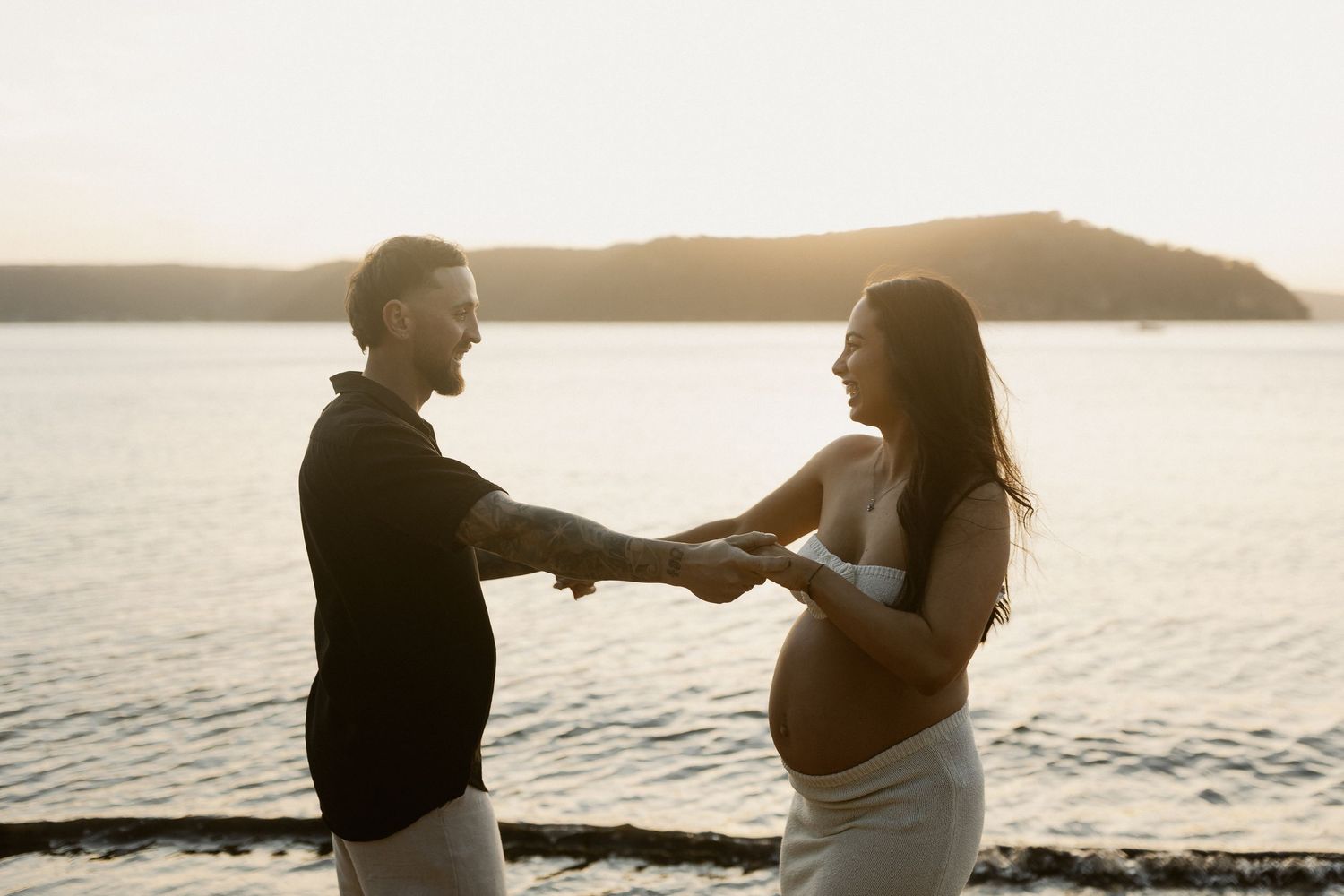 Maternity photoshoot captures couple in intimate pose by ocean during golden hour sunset.