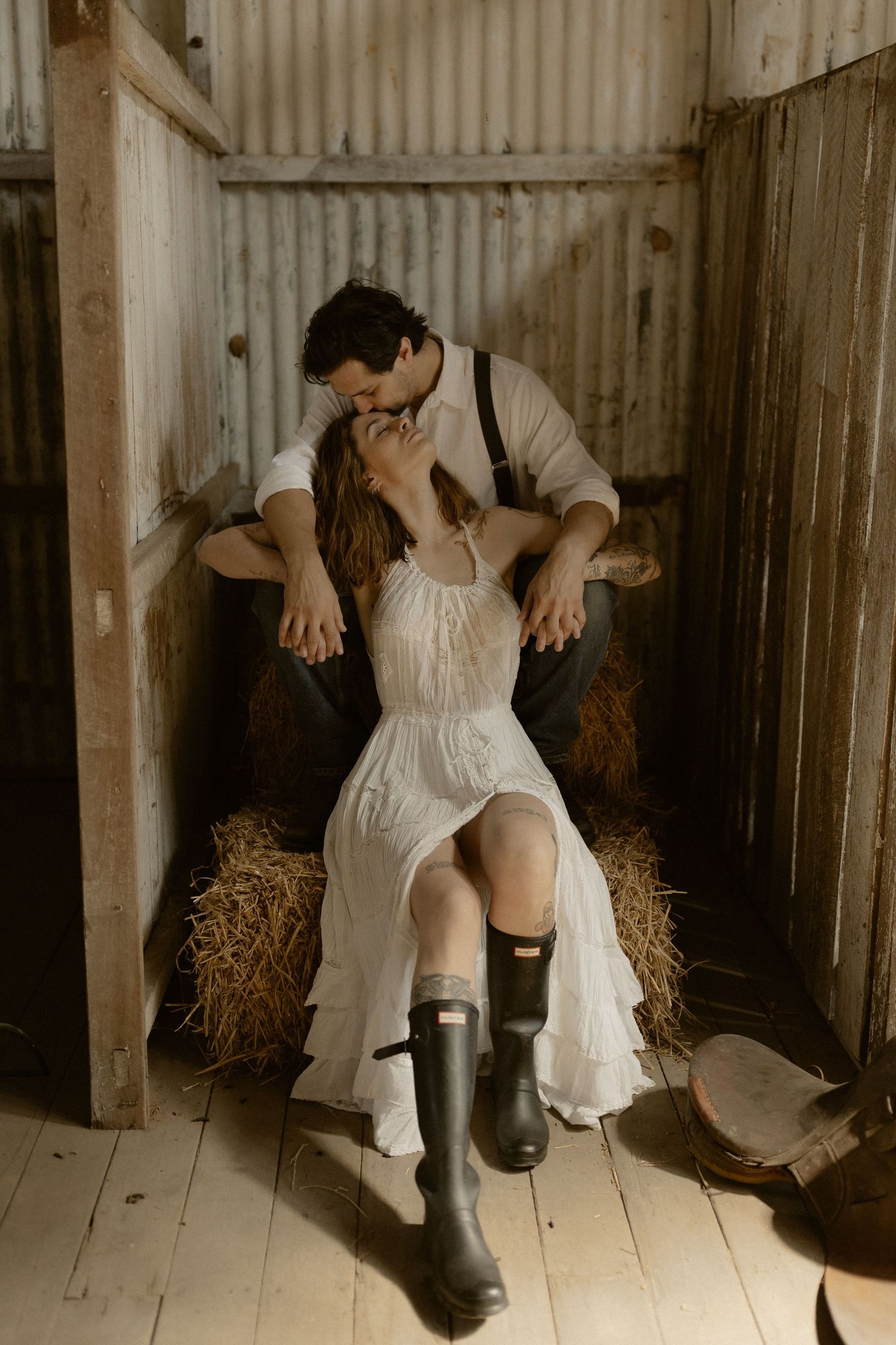 A couple sits romantically on hay bales inside a rustic barn setting.