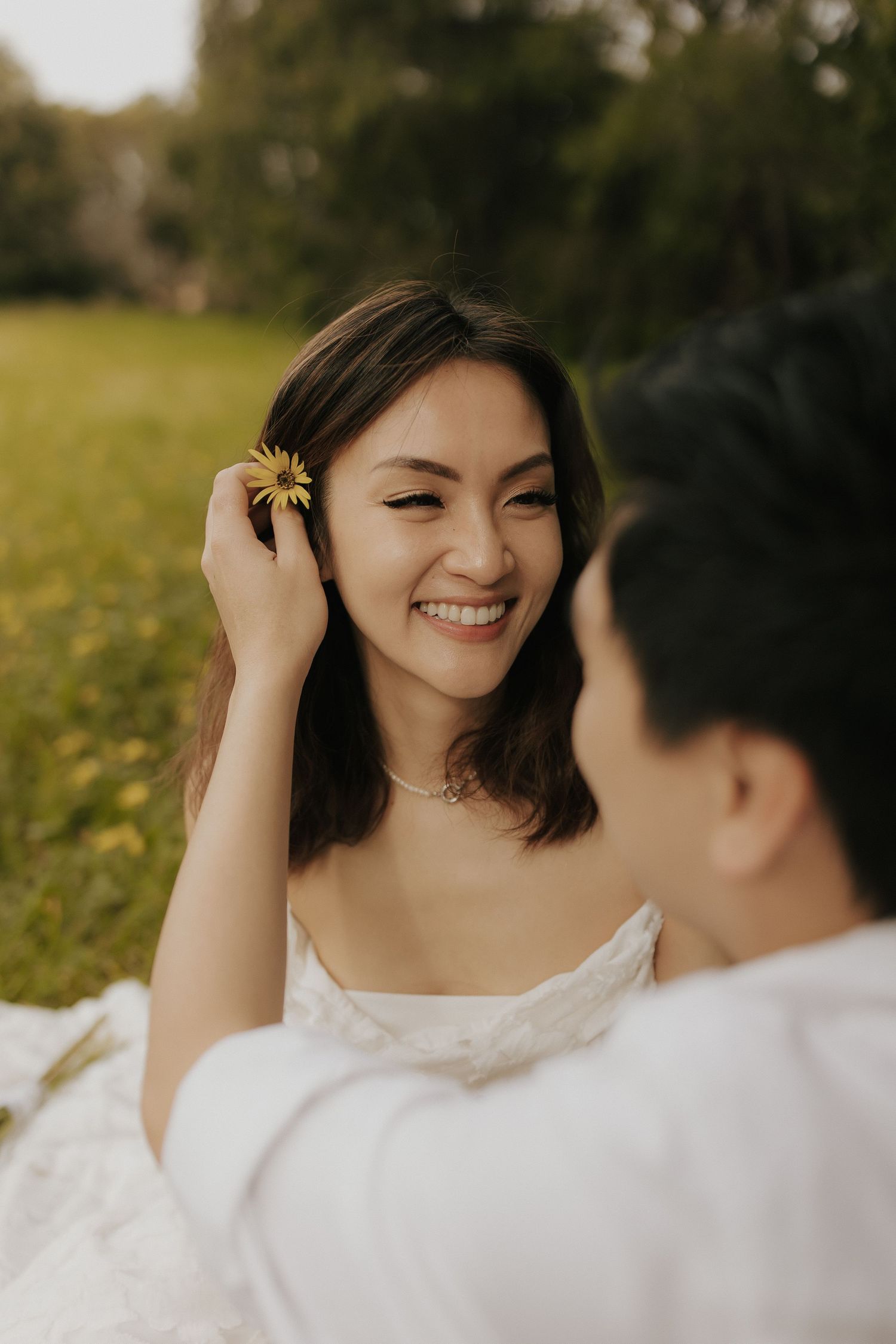 A joyful moment captured as a bride in white touches a yellow flower in her hair during an outdoor photoshoot.
