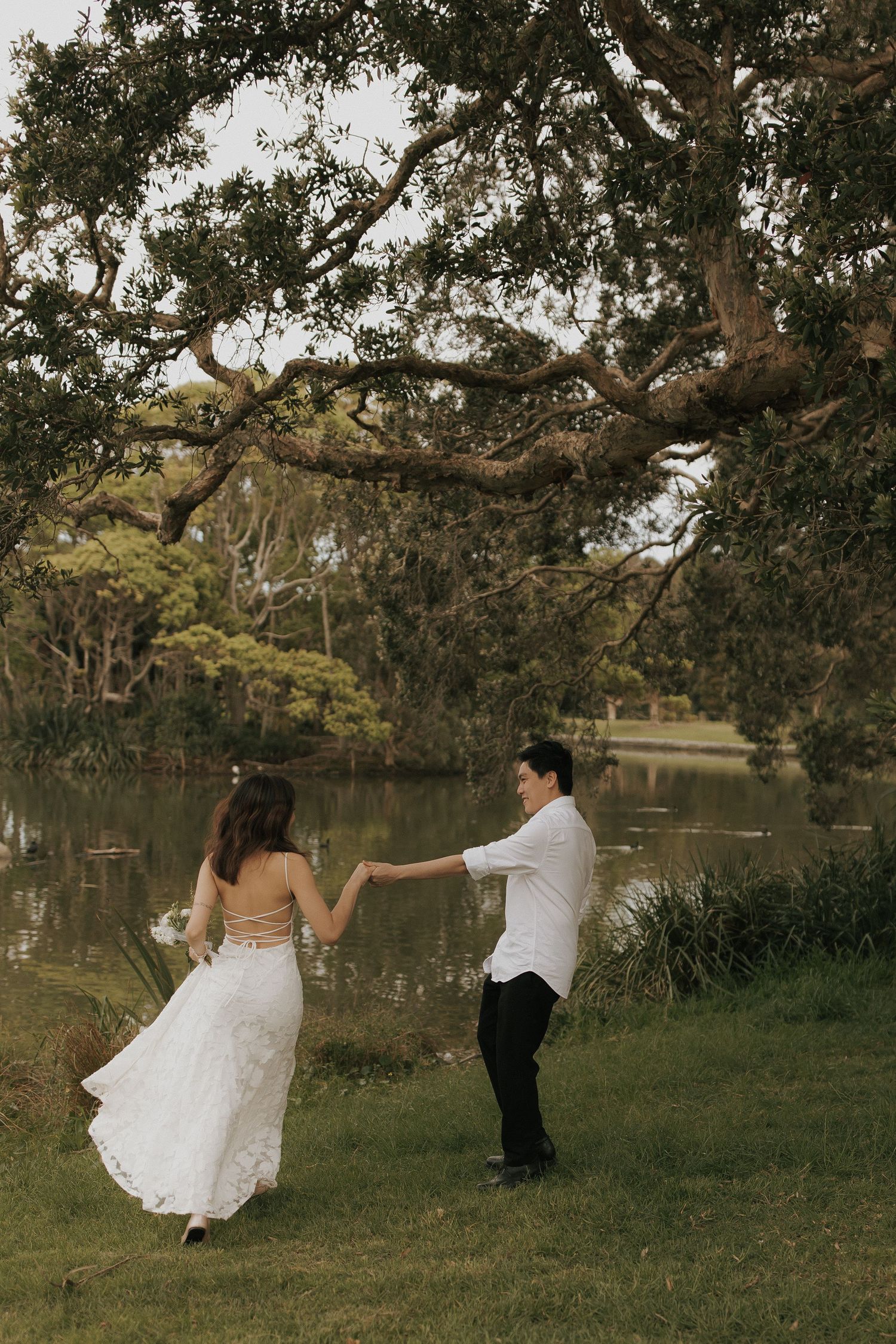 A romantic couple dances playfully under trees by a lake in an outdoor photoshoot.