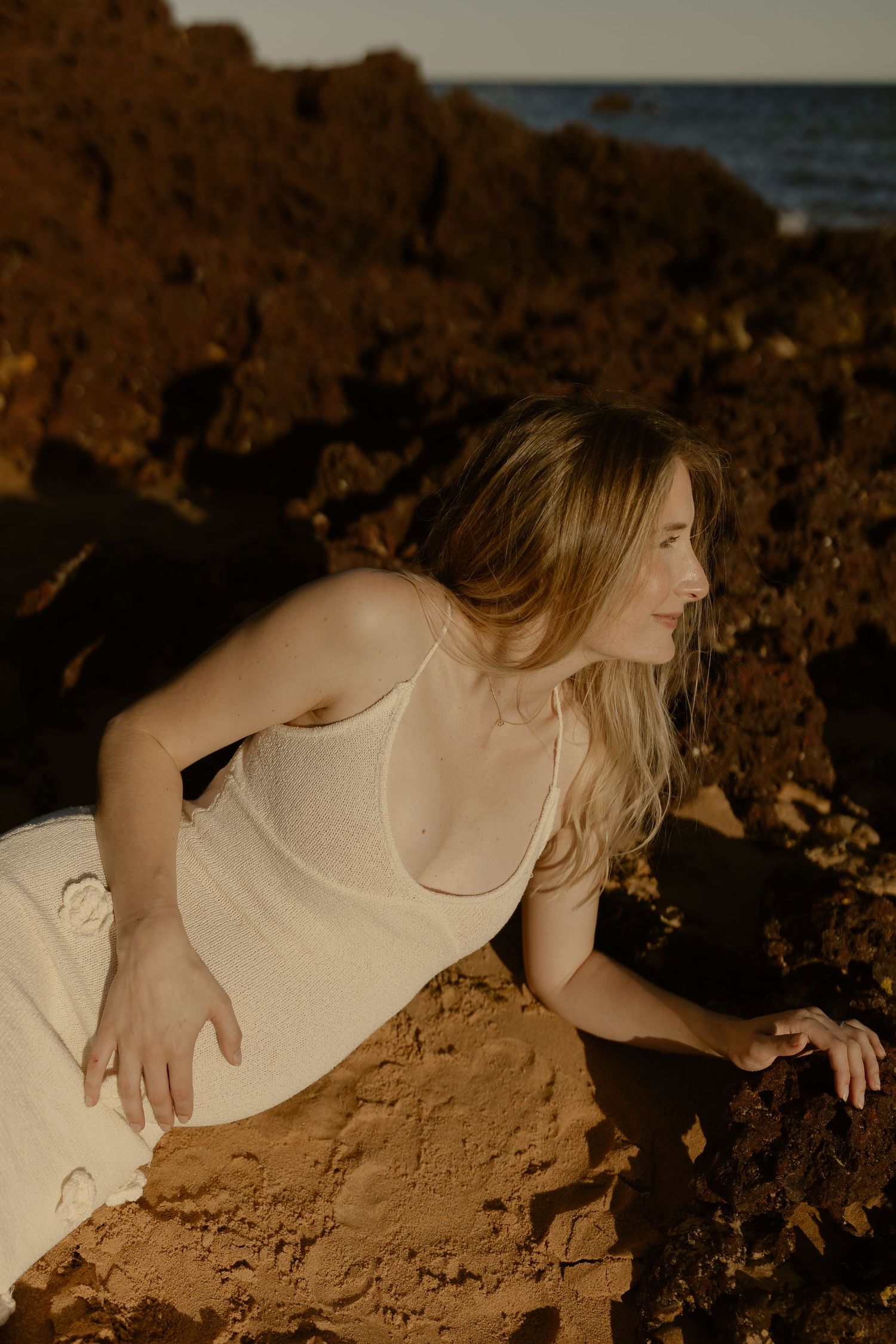 A dreamy golden-hour portrait on rugged coastal rocks features a cream-colored dress against the ocean backdrop.