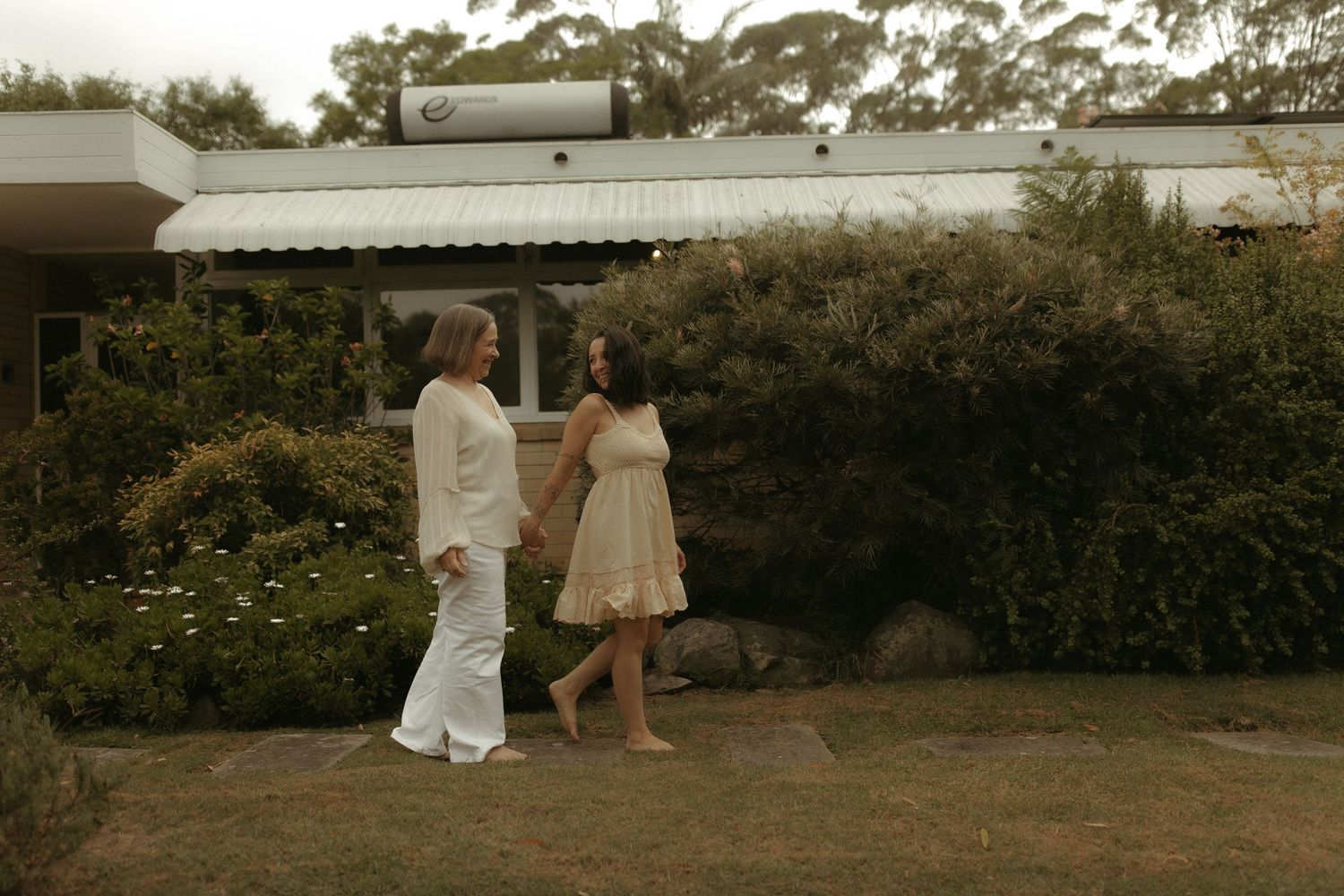 A couple walks together in a garden setting with modern architecture in the background.