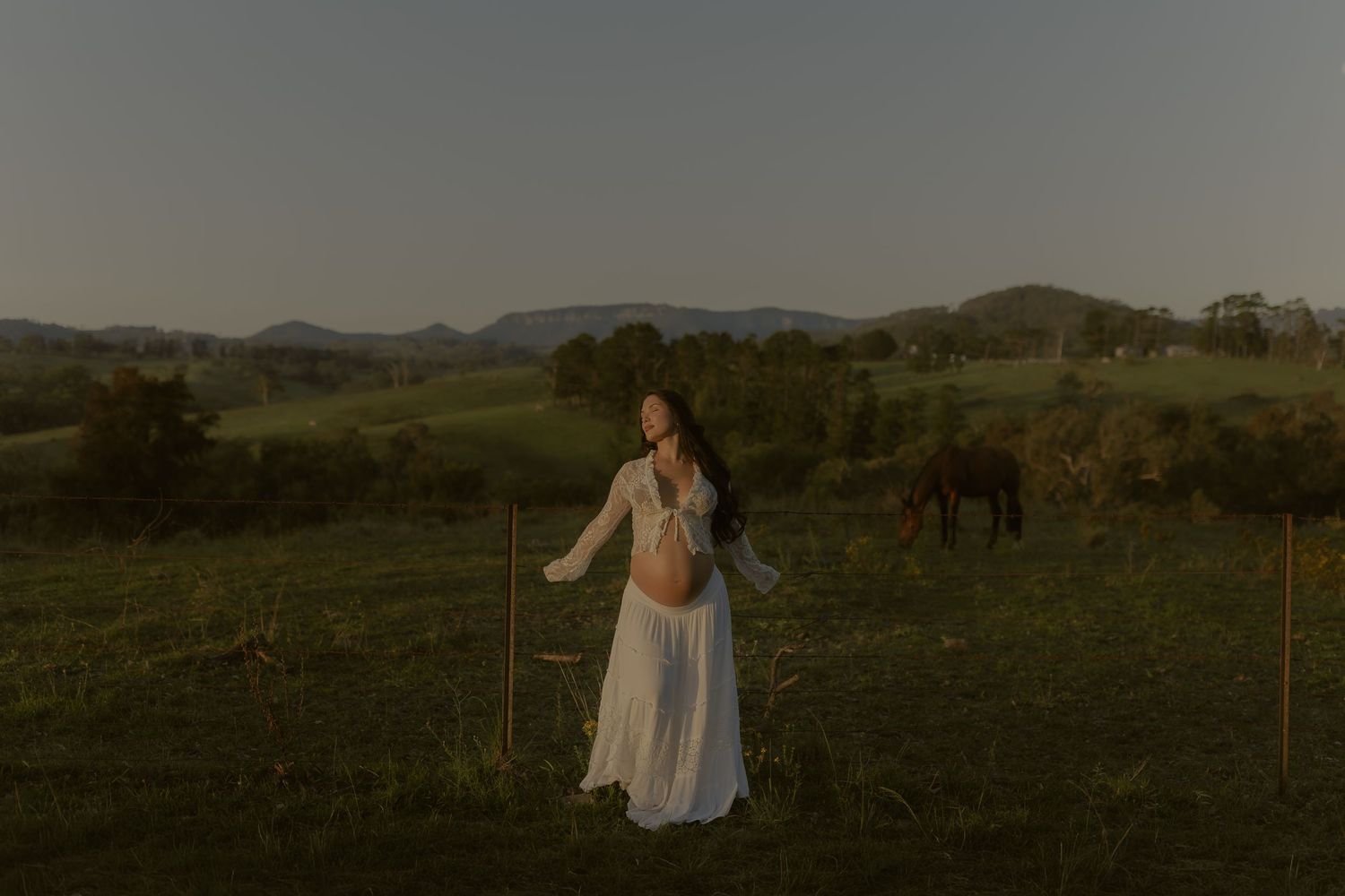 A figure in a white dress twirls gracefully in a scenic meadow with mountains in the background at dusk.