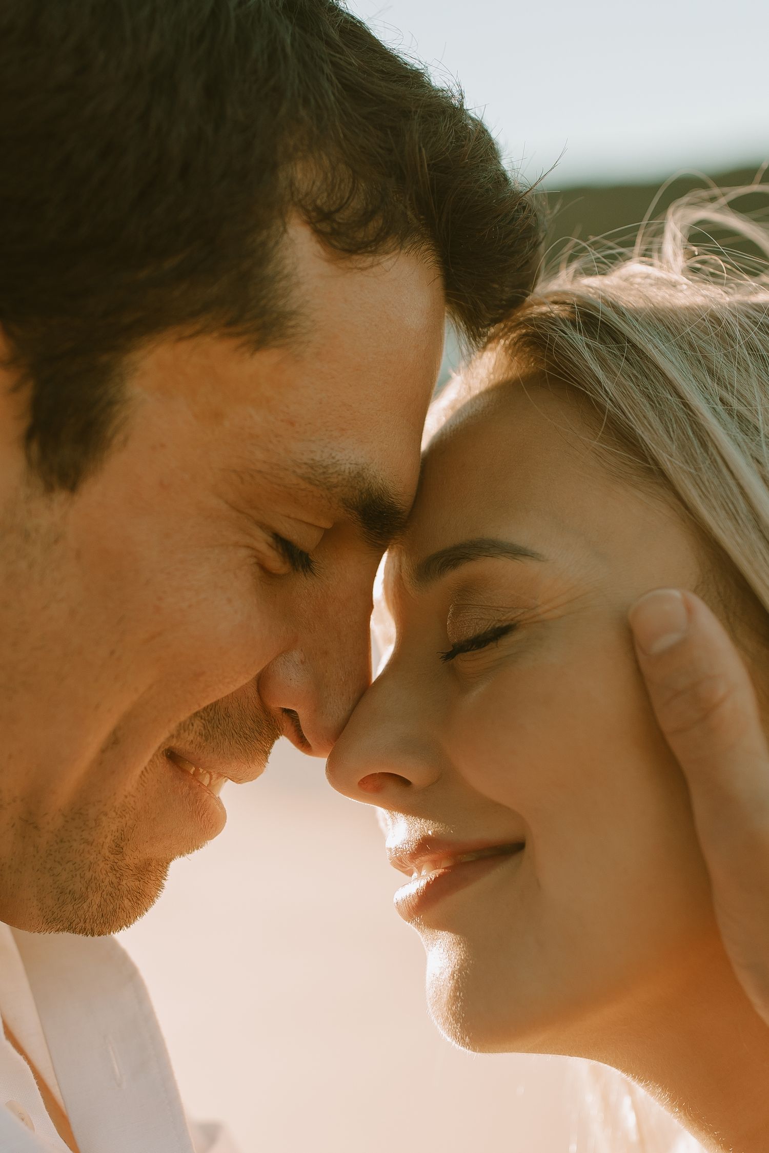 A romantic close-up photo of a couple touching foreheads while smiling in warm sunlight.
