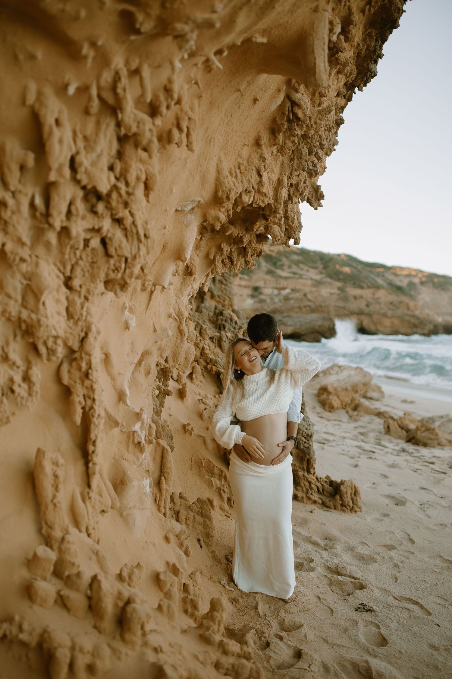 Person in white dress poses against textured rock formation at a scenic beach cove.