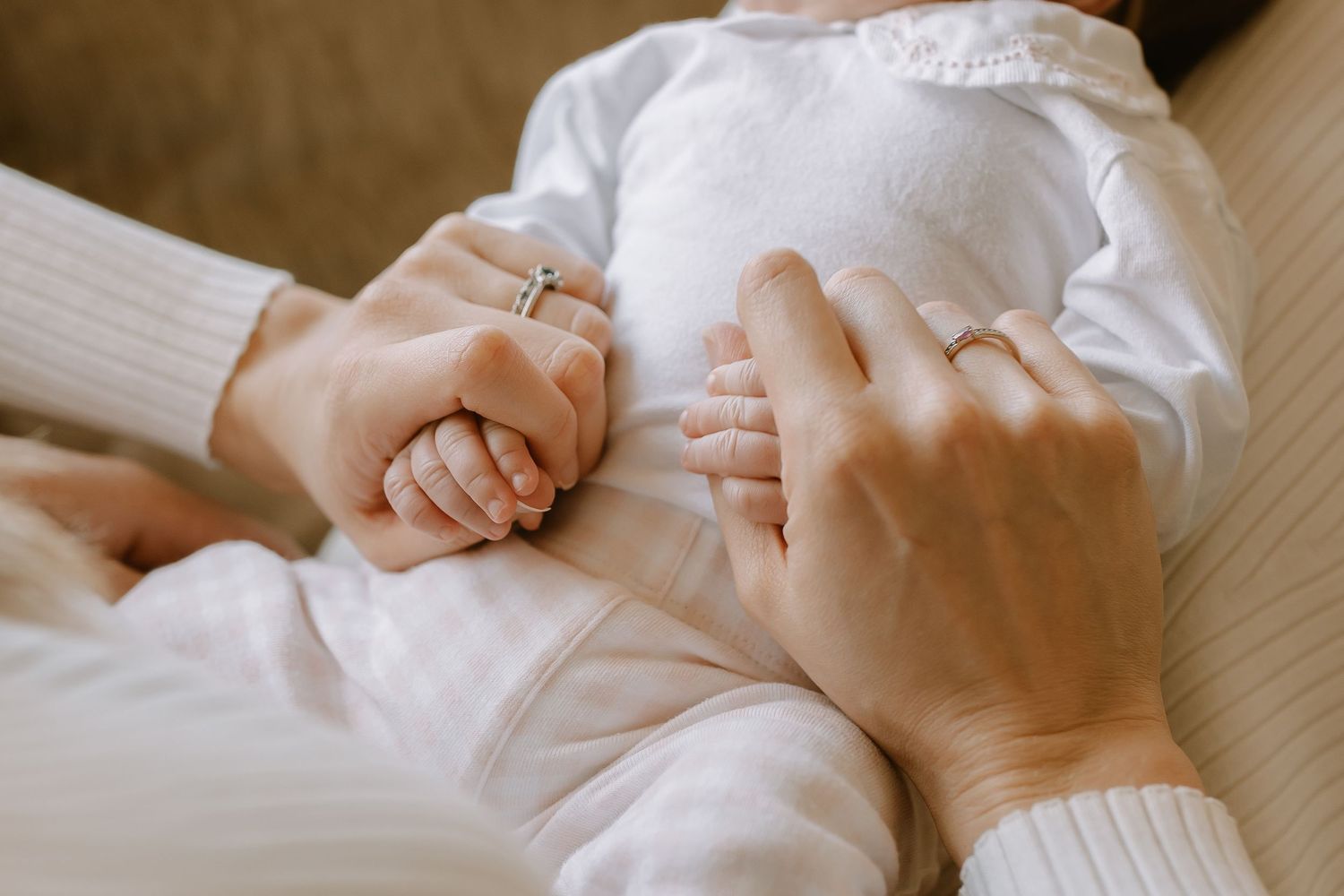 Two hands gently hold a baby wearing a white outfit, with a wedding ring visible on one hand.