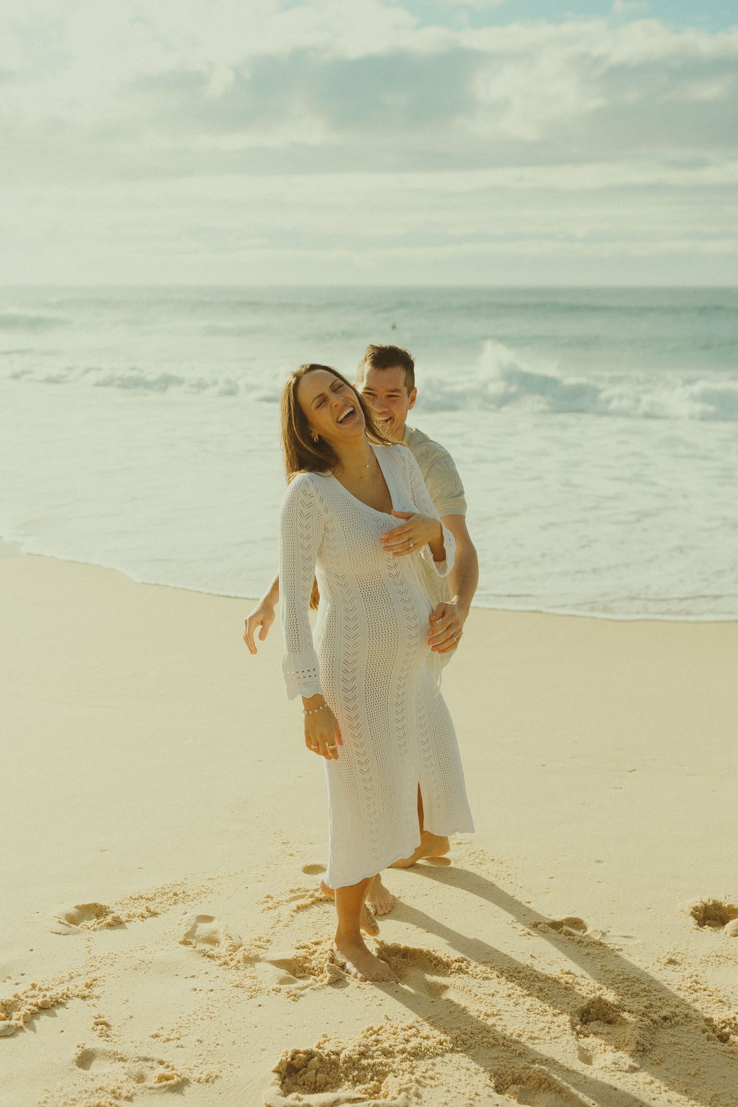 Two people share a joyful candid moment together on a sunny beach with crashing waves in the background.