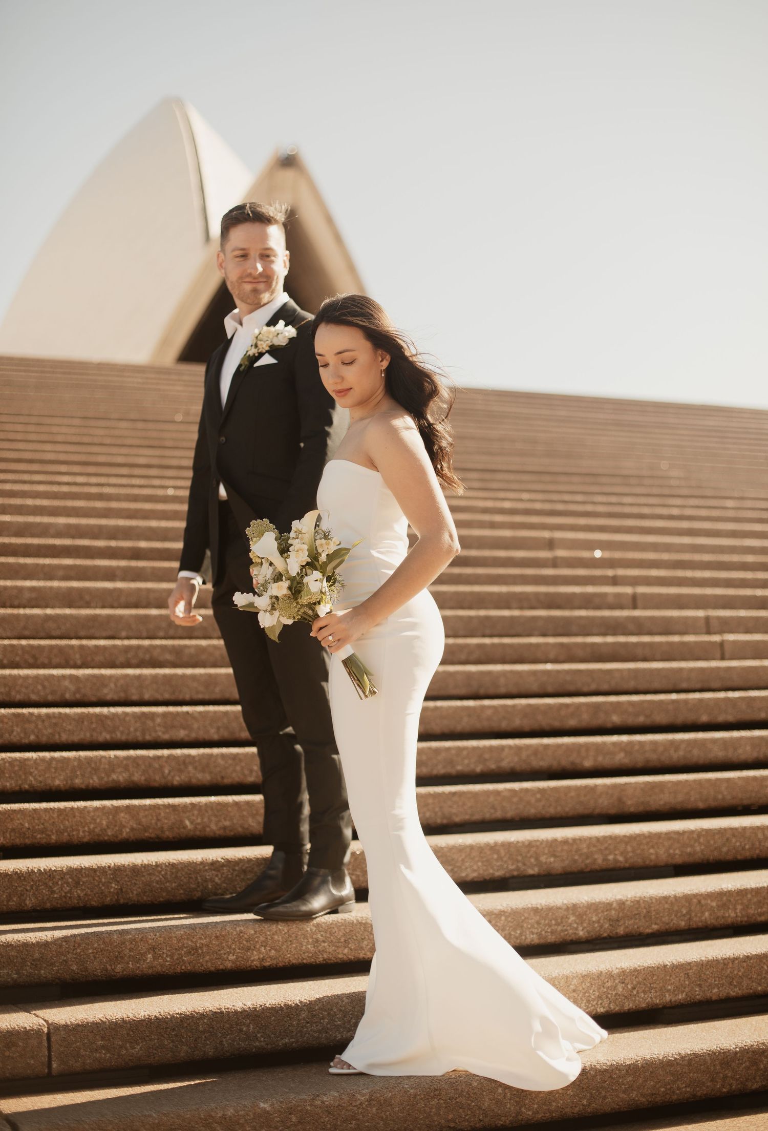 Bride with bouquet and groom stand on steps at Sydney Opera House in warm sunlight.