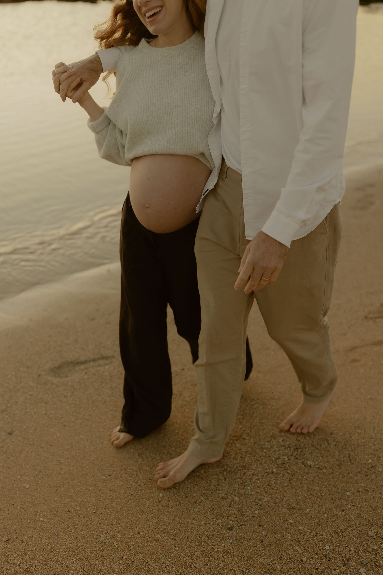 Pregnant couple walking barefoot on sandy beach at golden hour, woman showing baby bump in casual outfit.