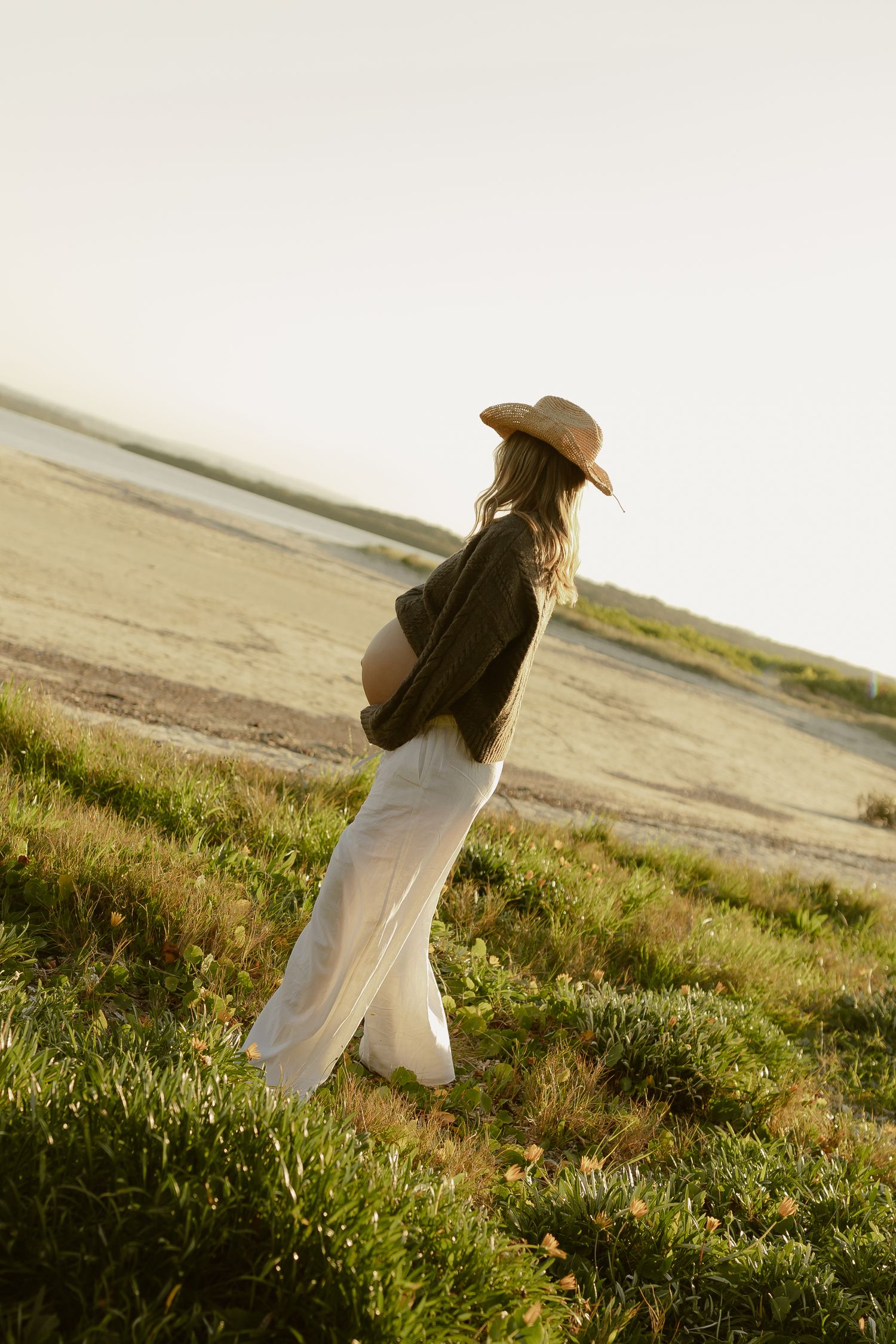 Pregnant woman in straw hat and white pants standing in golden field cradling her baby bump at sunset.