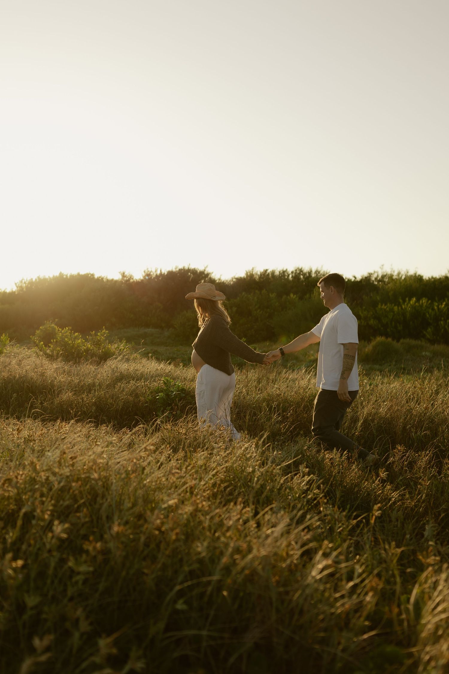Expecting couple holding hands while walking through tall grass field bathed in warm sunset light.