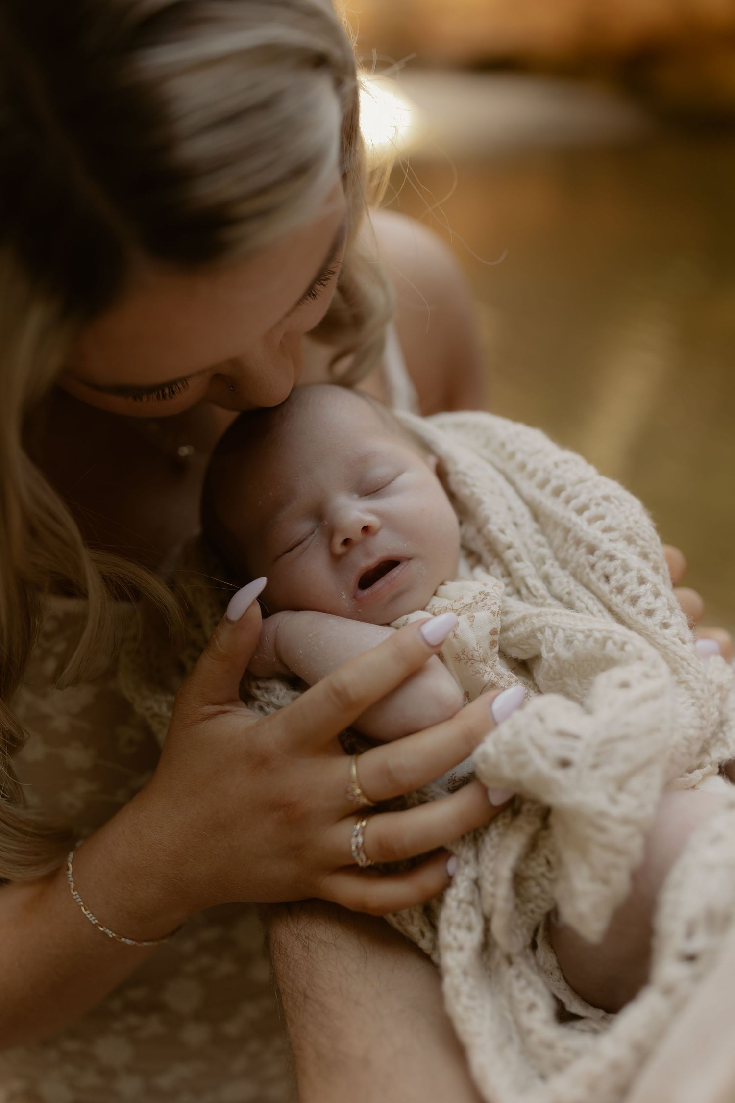 A mother tenderly holds her sleeping newborn wrapped in a cream crochet blanket, leaning in for a gentle kiss.