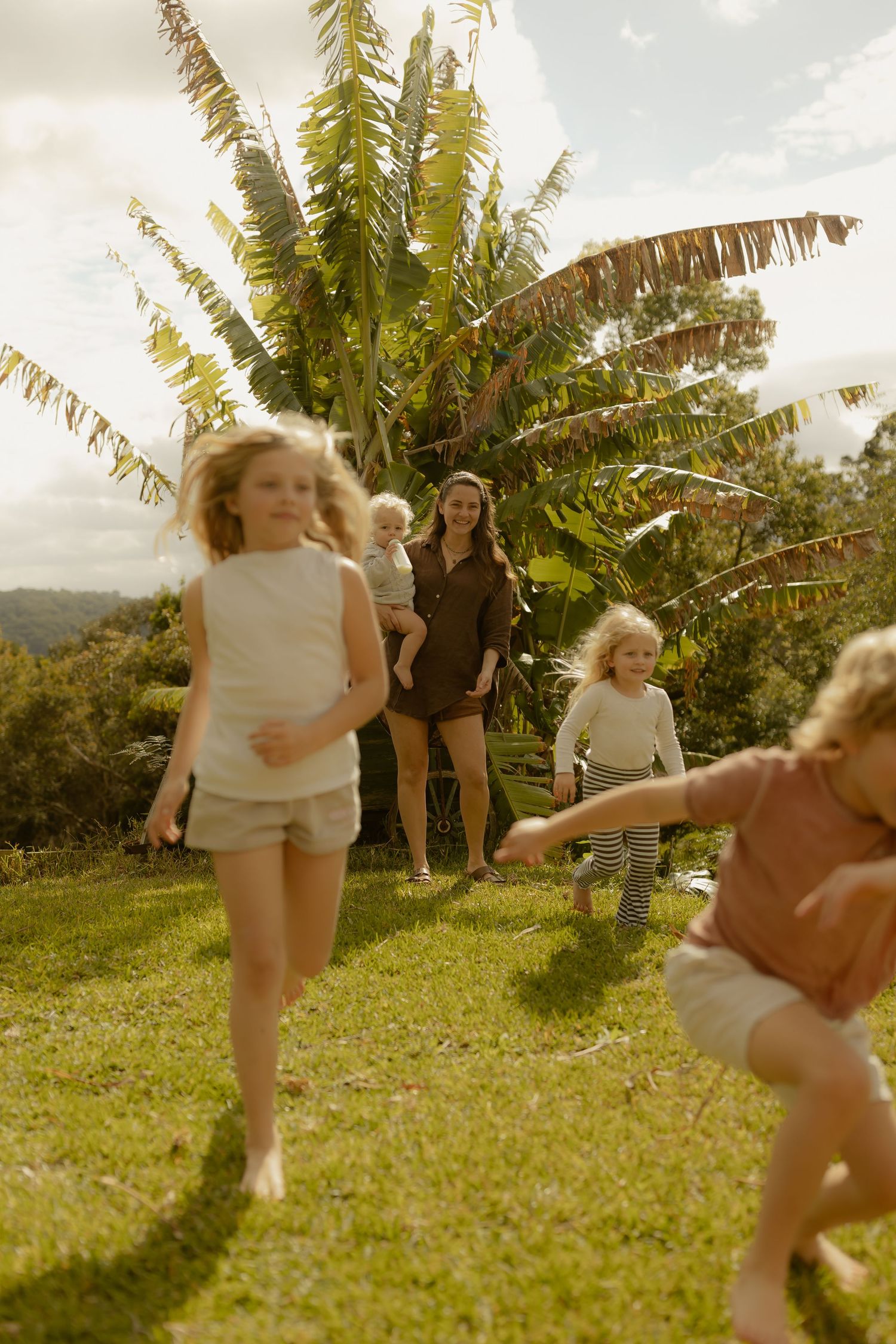 Children playing on green grass outdoors with a woman holding a baby near tropical plants on a sunny day.