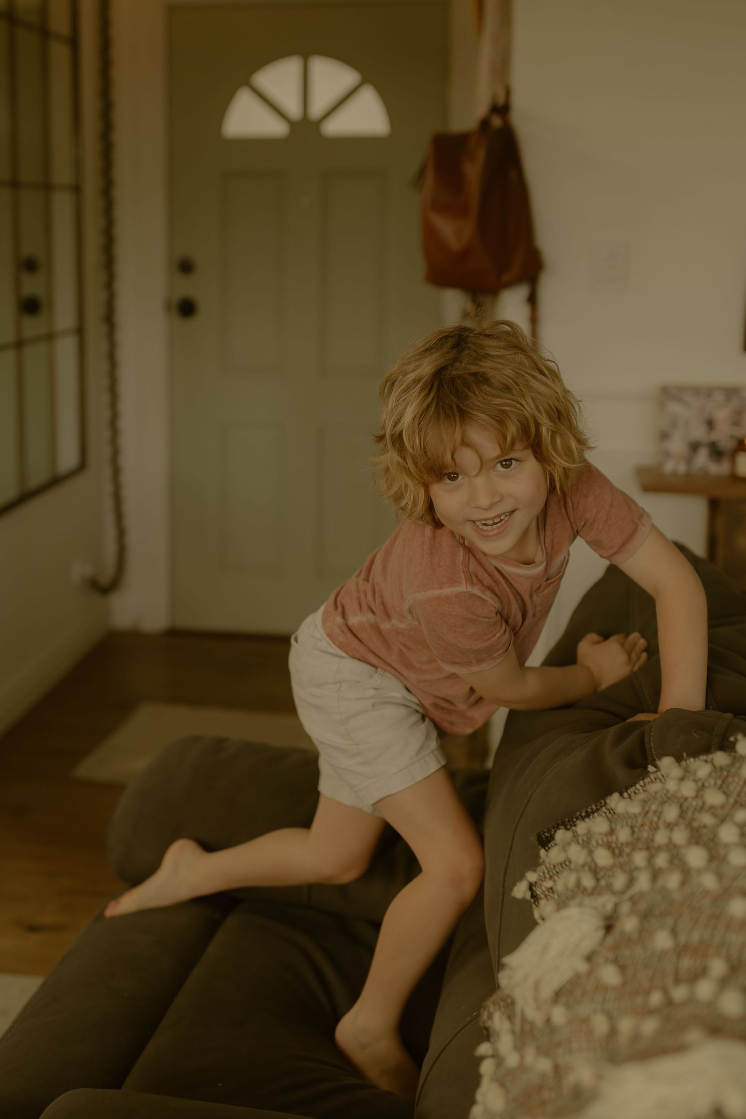 A young barefoot boy with wavy hair climbs over a dark sofa, smiling playfully in a cozy home interior.