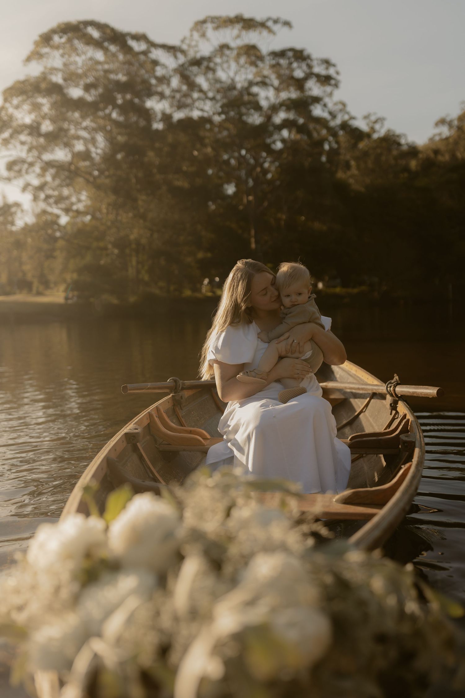 Mother kissing baby in wooden rowboat on calm lake at golden hour, white flowers in foreground.
