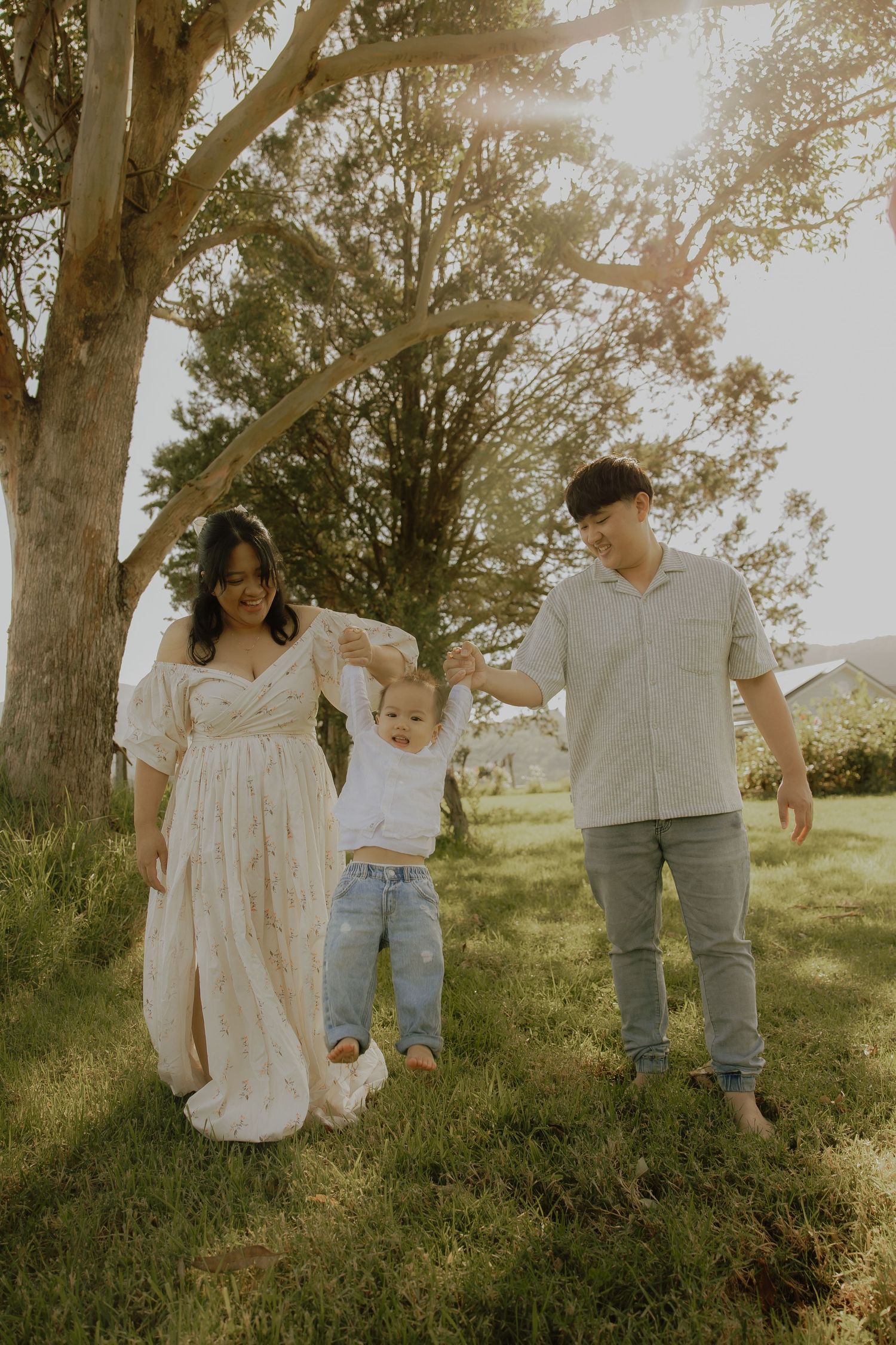 Young family holding toddler's hands under sunlit tree on green grass, casual outdoor portrait.