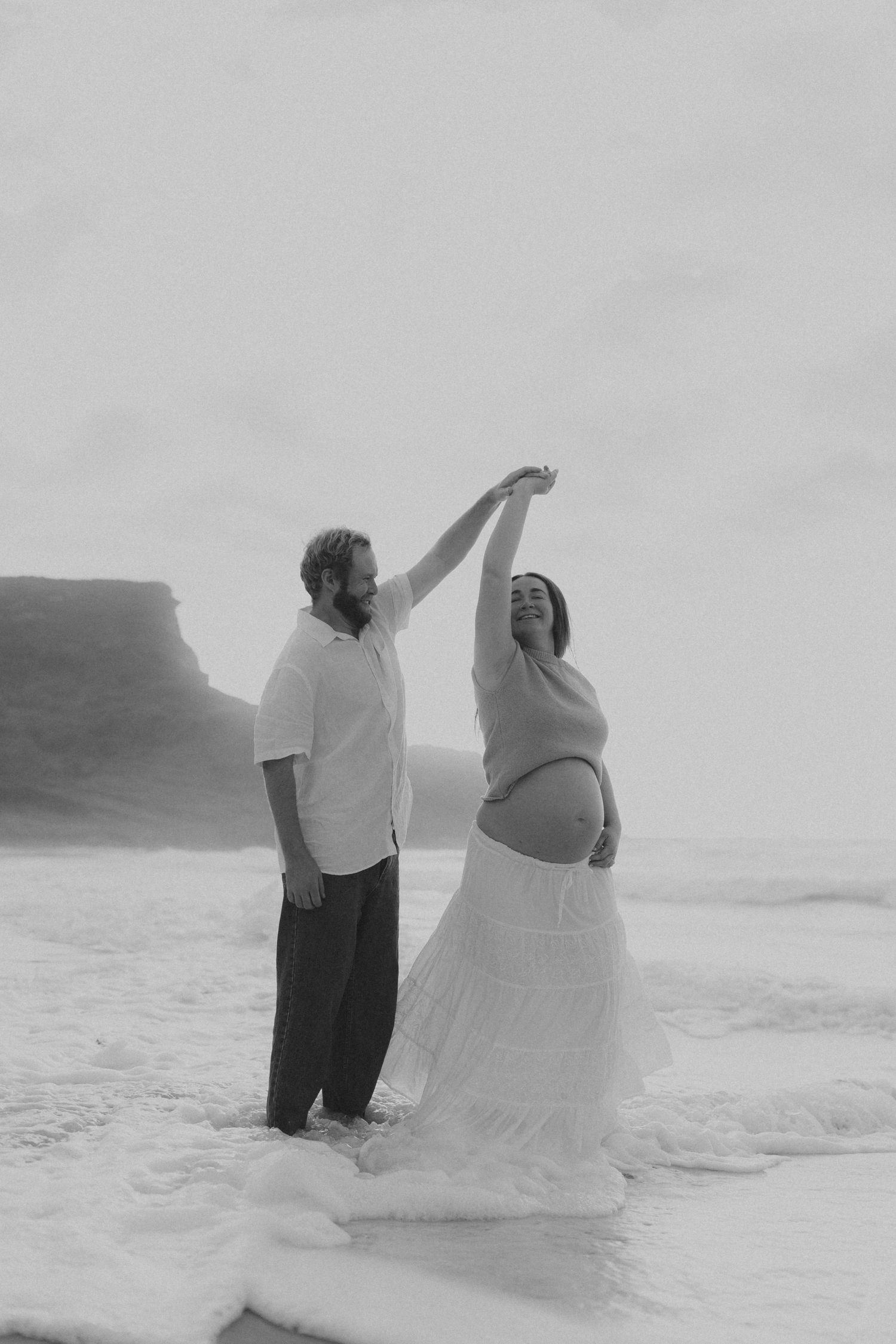 A couple dances together on a misty beach with dramatic coastal cliffs in the background.