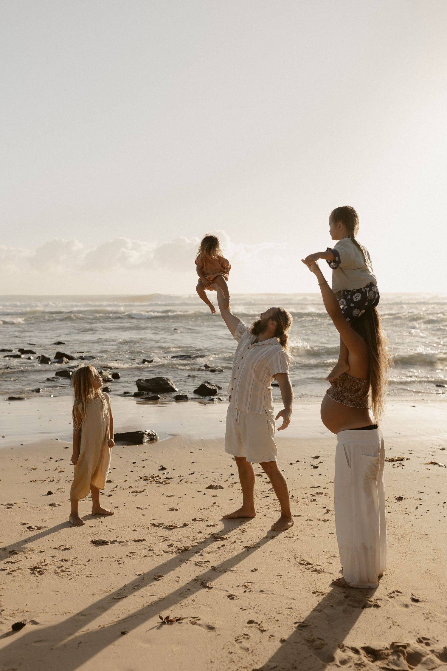 Three people in white dresses walk along a rocky beach at sunset casting long shadows on the sand.