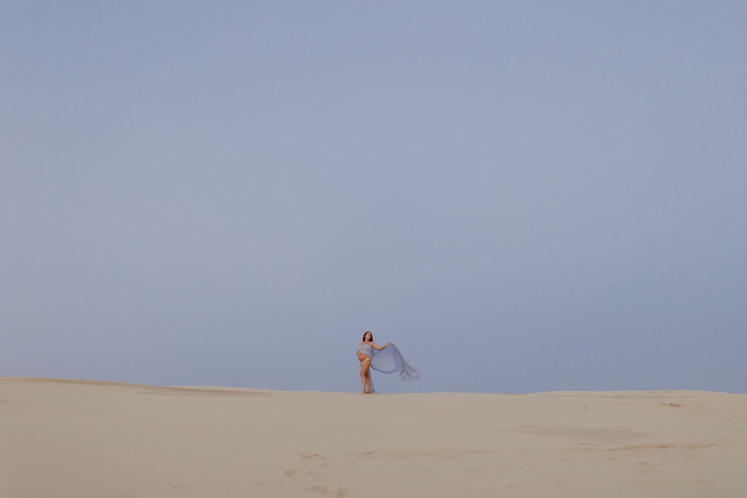 Distant silhouette with flowing fabric stands alone on vast desert dunes against blue sky.