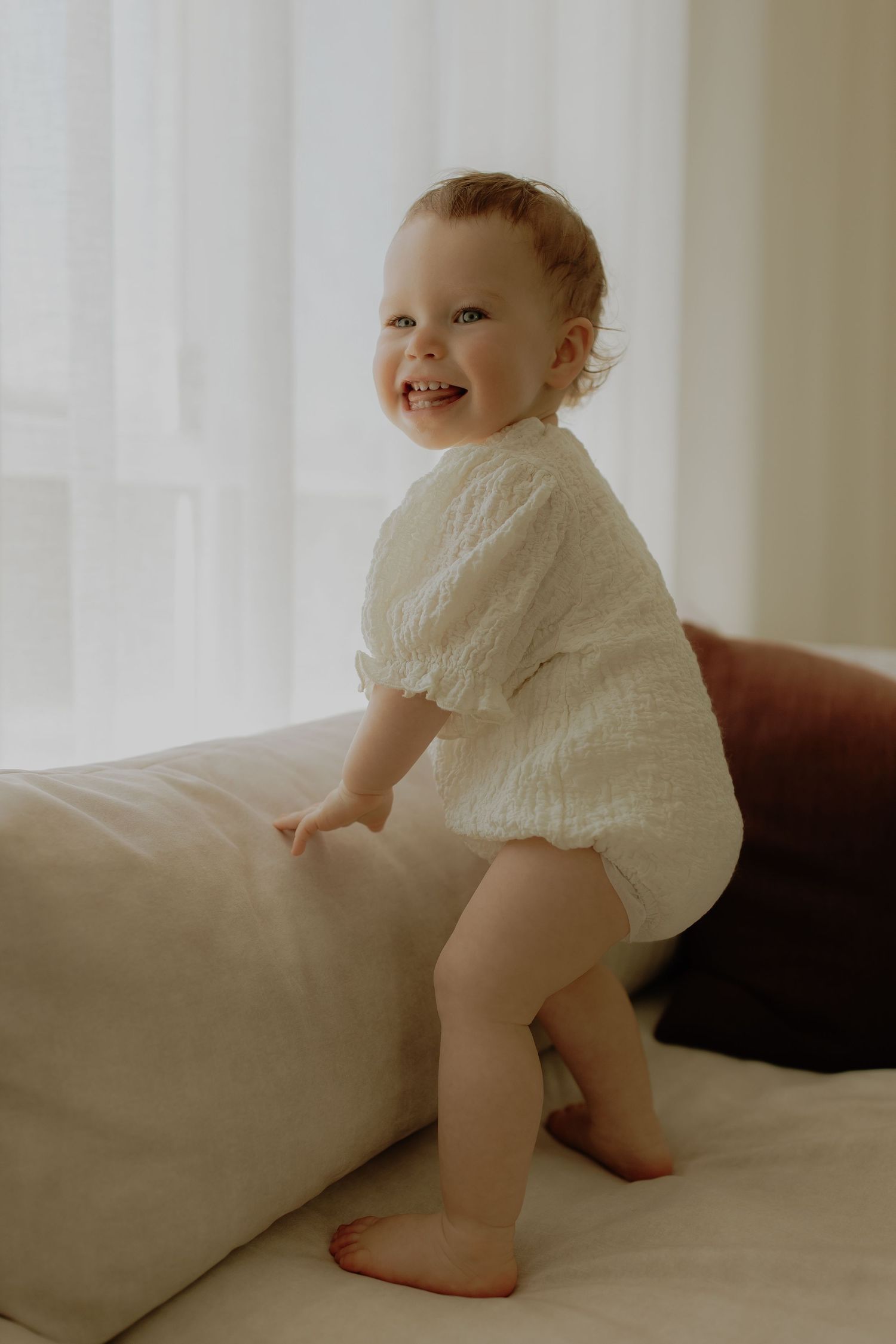 Smiling baby in white lace romper standing on a cream sofa near a bright window.