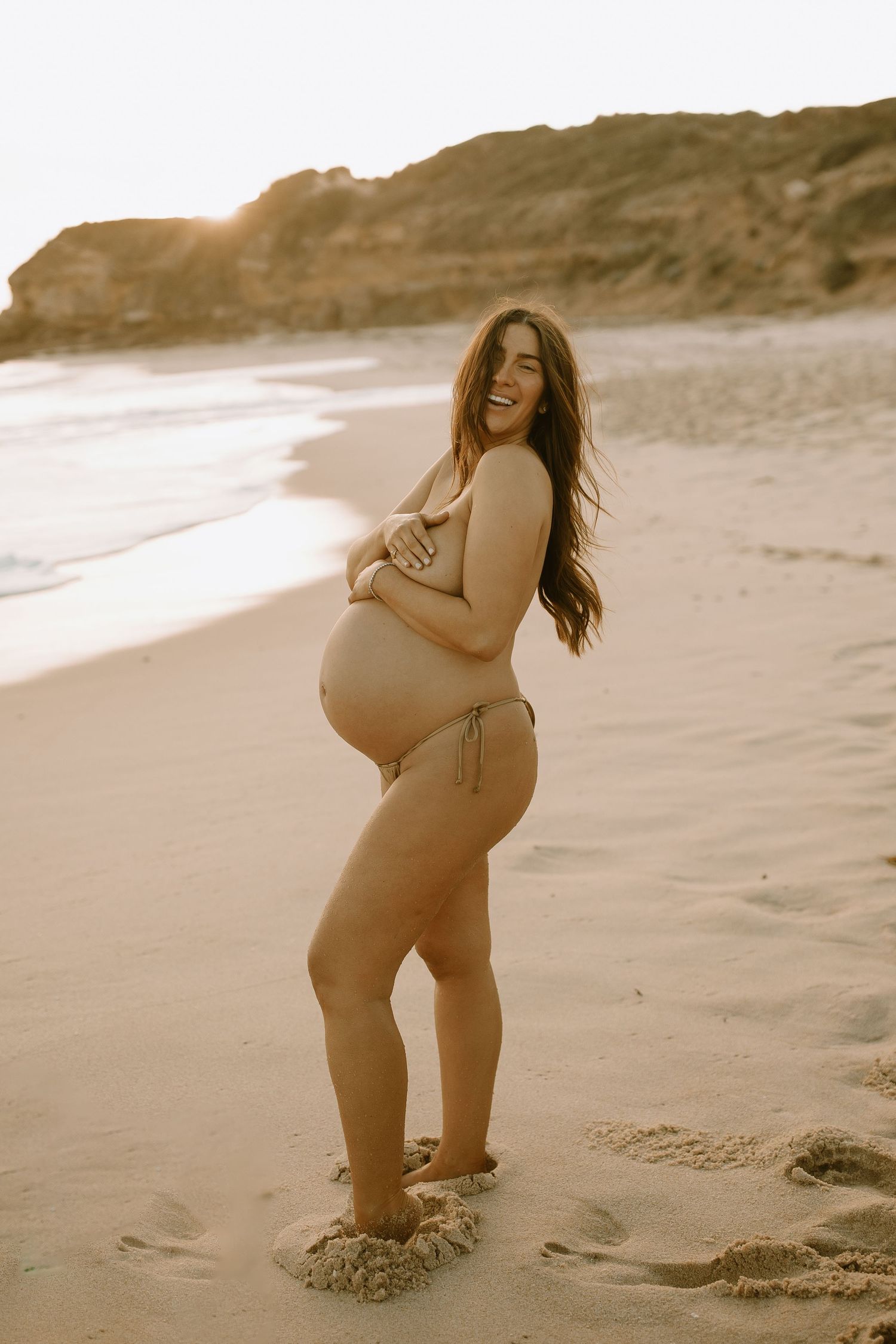 Expecting mother poses joyfully at sunset on a sandy beach with cliffs in the background.