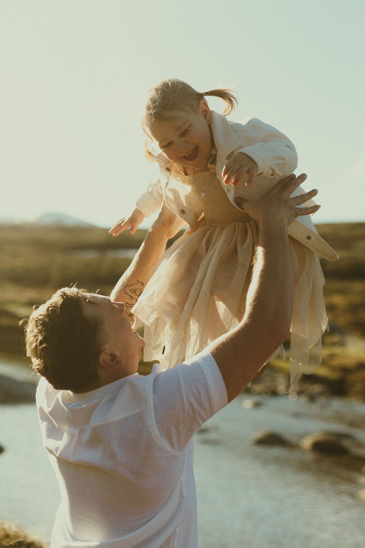 A parent lifts a child wearing a flowing dress against a golden sunset backdrop in an outdoor setting.