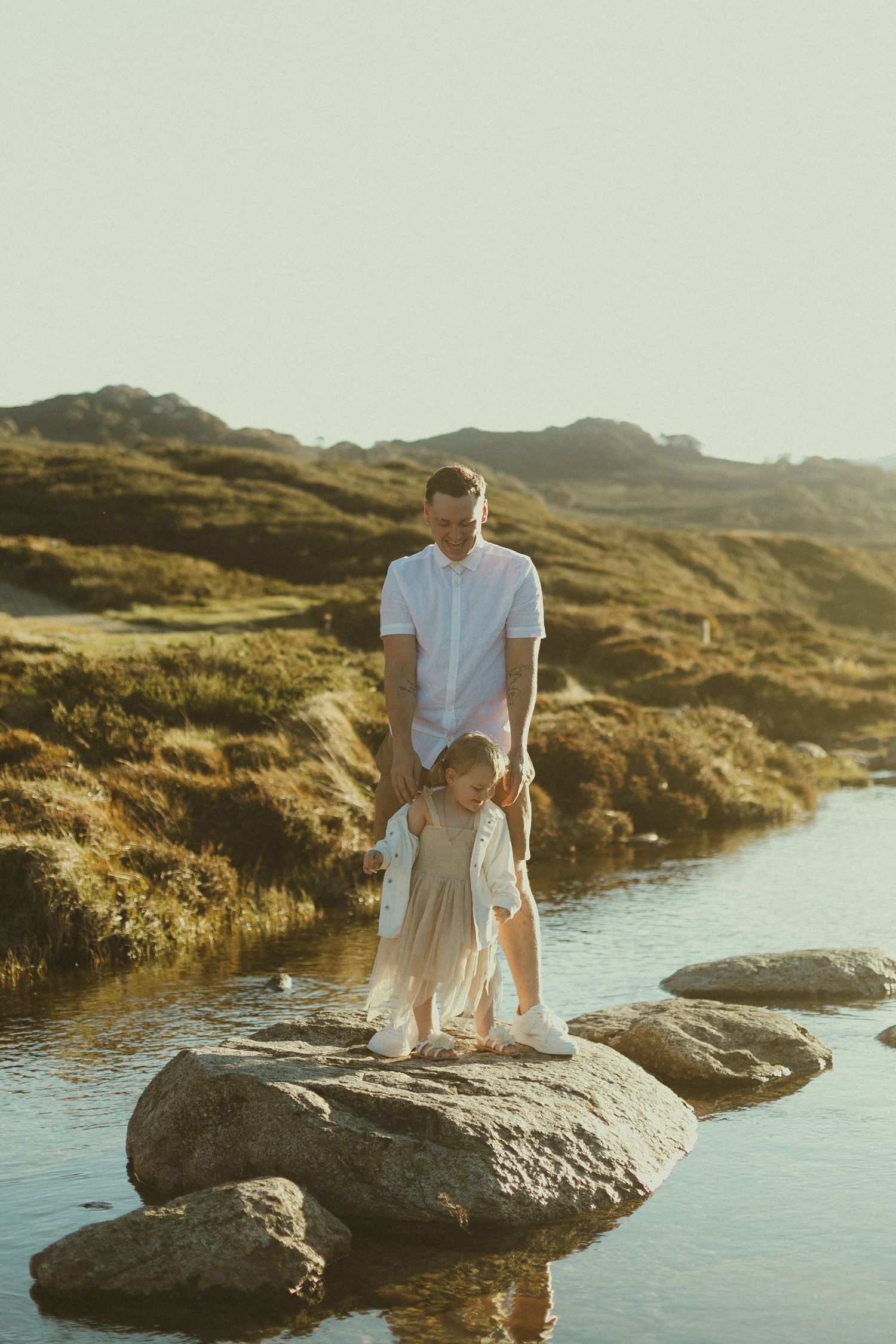 Two people stand on a boulder by a scenic coastal stream at sunset among rugged hills and grassy terrain.