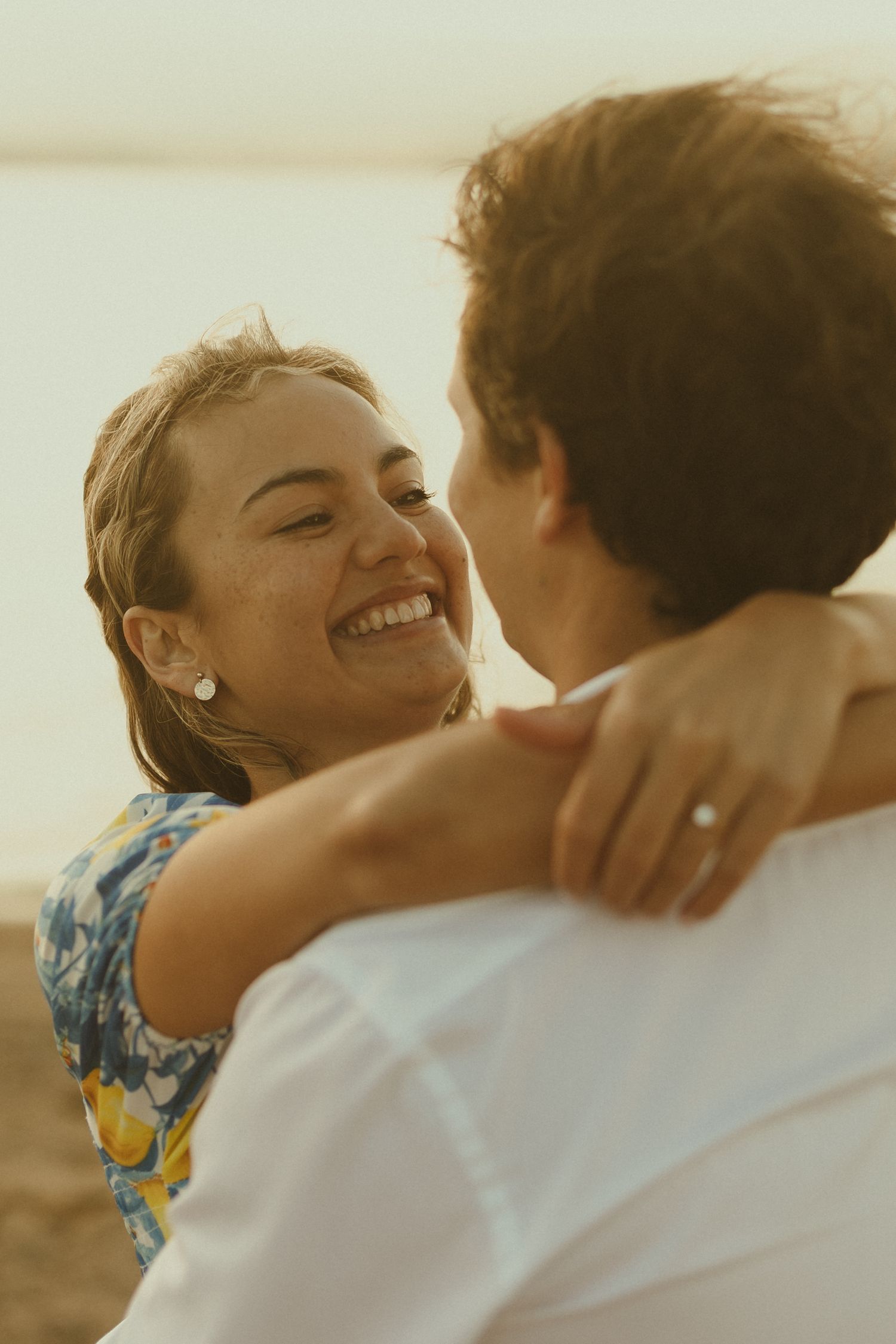 A romantic couple embraces during golden hour, with joyful expressions and a blue and yellow floral dress.